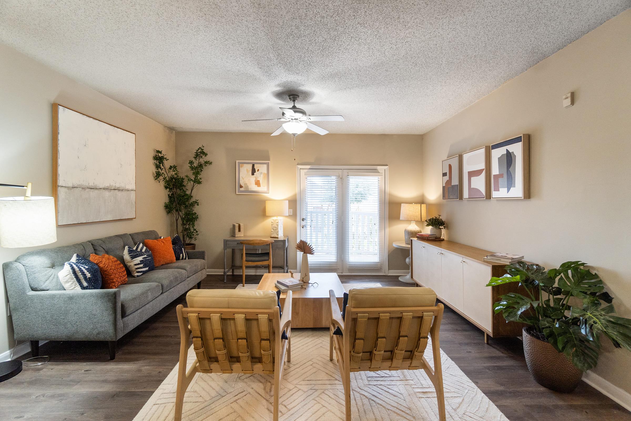 A bright and inviting living room featuring a gray sofa with colorful cushions, two wooden armchairs, a coffee table, and a lamp. Natural light streams in from sliding glass doors, and a potted plant adds greenery. Simple artwork decorates the walls, enhancing the cozy atmosphere.