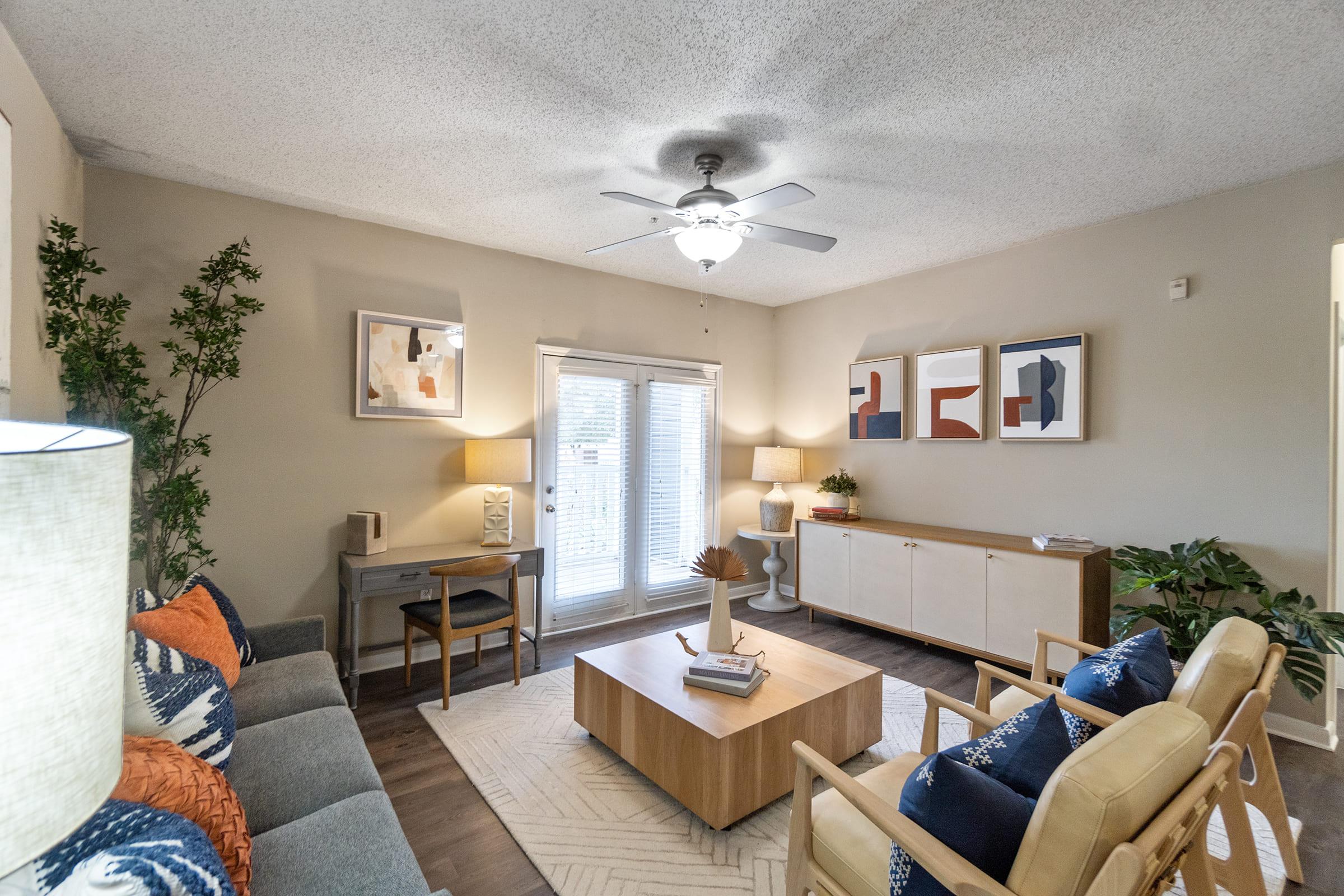 A cozy living room featuring a ceiling fan, a gray sofa with colorful cushions, and two stylish chairs. There’s a wooden coffee table in the center, a console table against the wall, and a large window with blinds. Decor includes framed artwork and potted plants, creating a welcoming atmosphere.