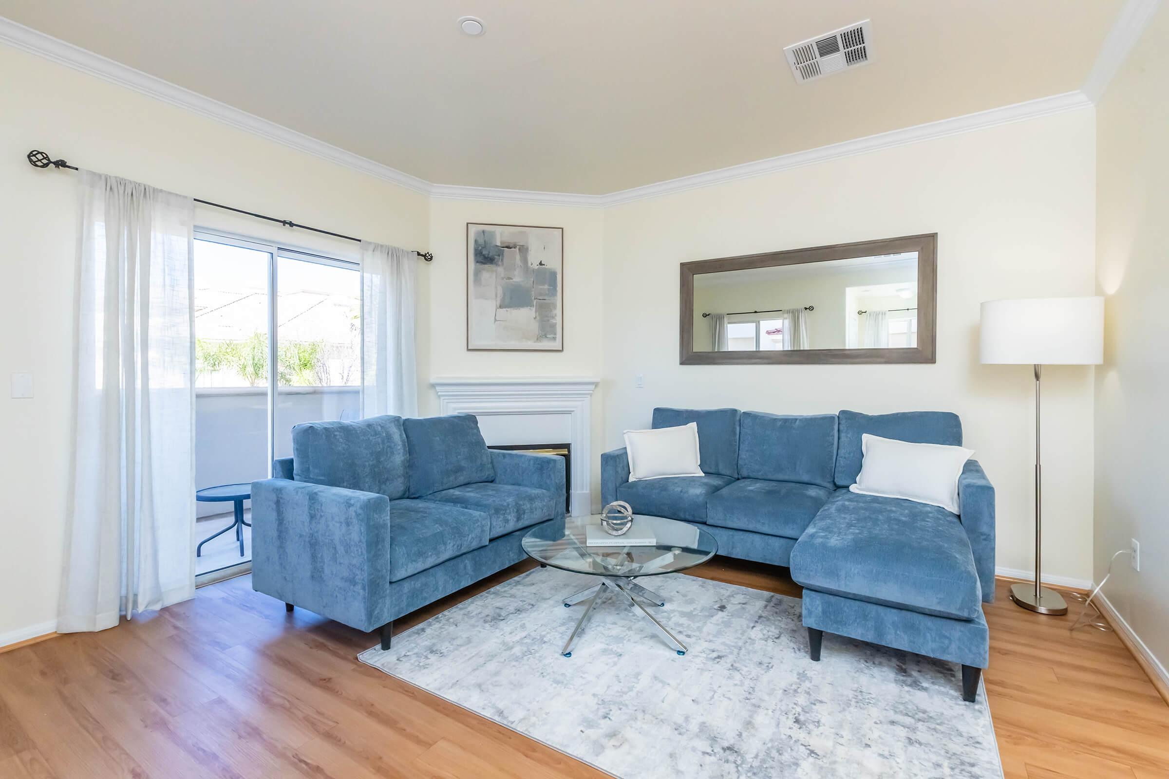 Cozy living room featuring a blue sectional sofa and a matching loveseat, a glass coffee table in front, and a fireplace. Natural light streams in through sheer curtains, and a large mirror reflects the space. A simple artwork adorns the wall, and a lamp stands in the corner, enhancing the inviting atmosphere.