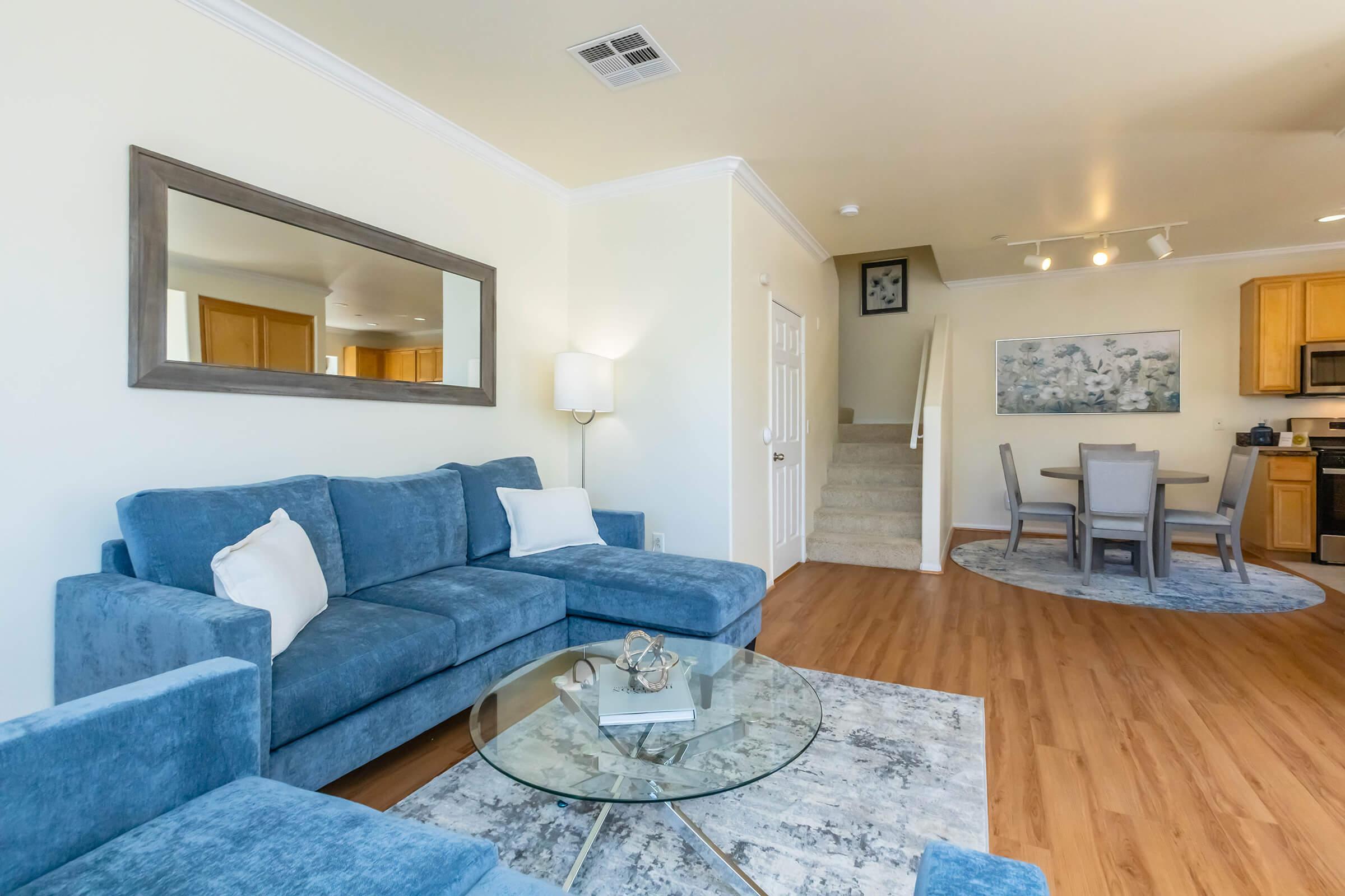 Bright living room featuring a blue sectional sofa with white pillows, a round glass coffee table, and a modern area rug. There is a staircase leading to another level, a dining table with chairs in the background, and a large mirror on the wall. Natural light illuminates the space, creating a welcoming atmosphere.