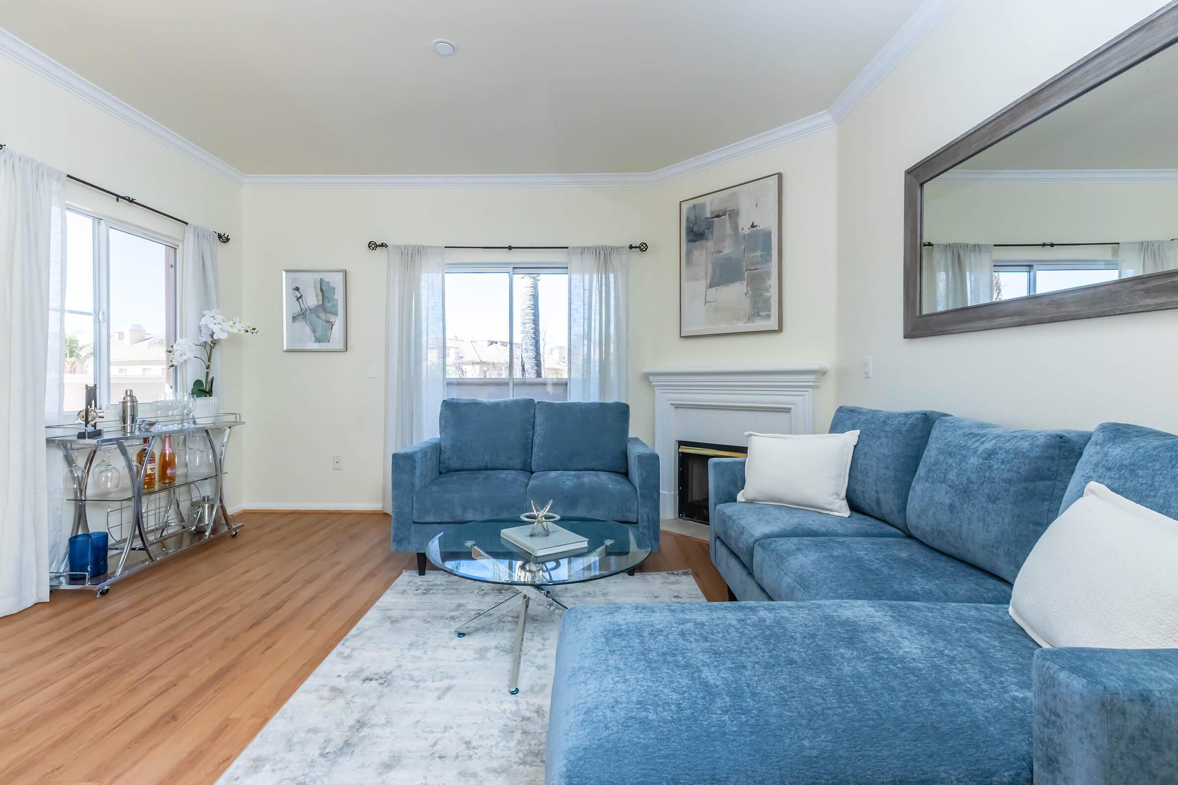 A bright and airy living room featuring a blue sectional sofa, a glass coffee table, and a decorative vase. Large windows allow natural light, complemented by sheer curtains. A corner fireplace and a wall mirror enhance the space's openness, while a light-colored area rug adds warmth to the wooden flooring.
