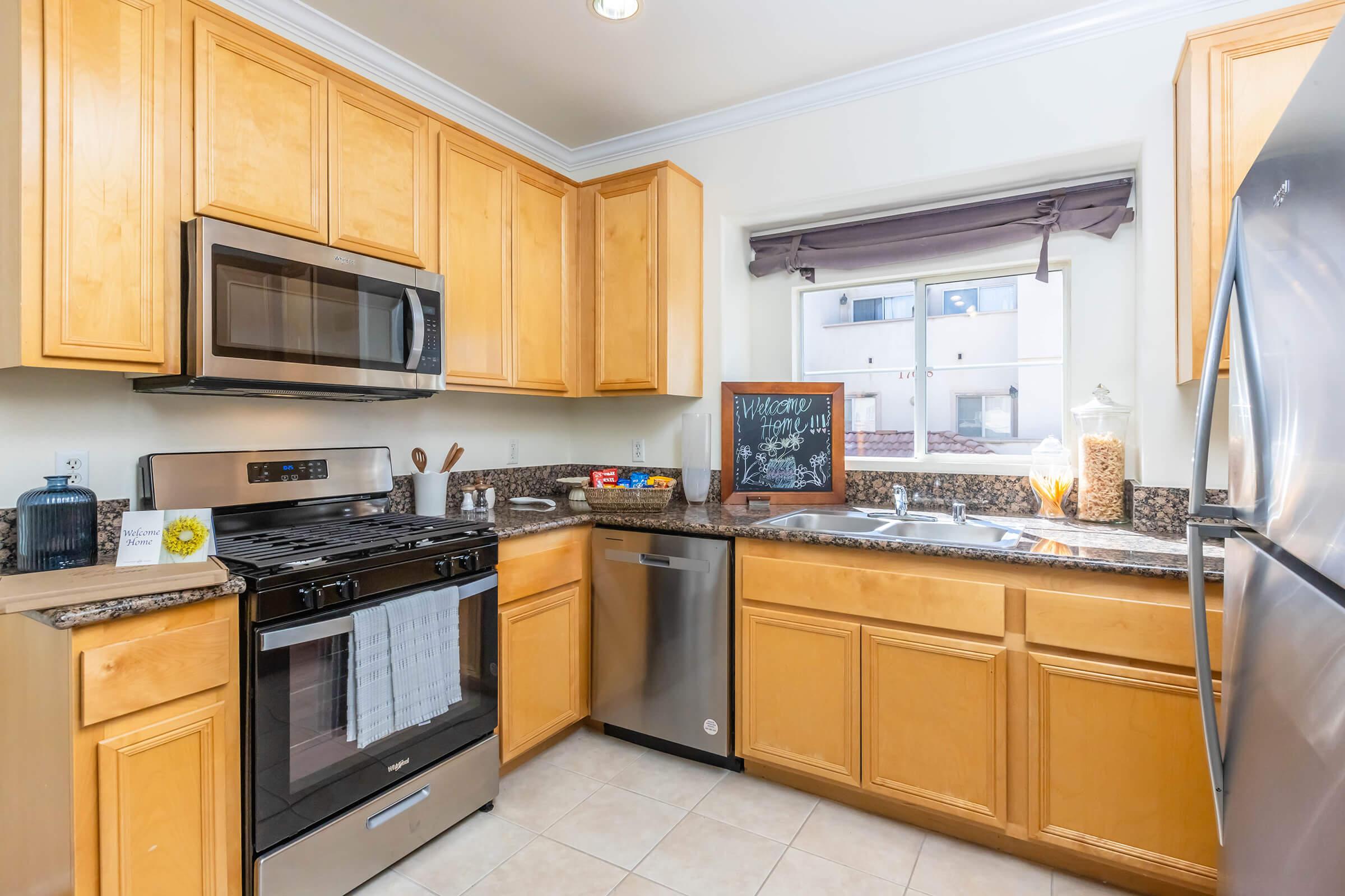 A modern kitchen featuring wooden cabinetry and granite countertops. The appliances include a stainless steel oven, microwave, and dishwasher. A window offers natural light, and a chalkboard on the counter displays a welcoming message. Several cooking utensils and a jar of cereal are visible on the countertops.