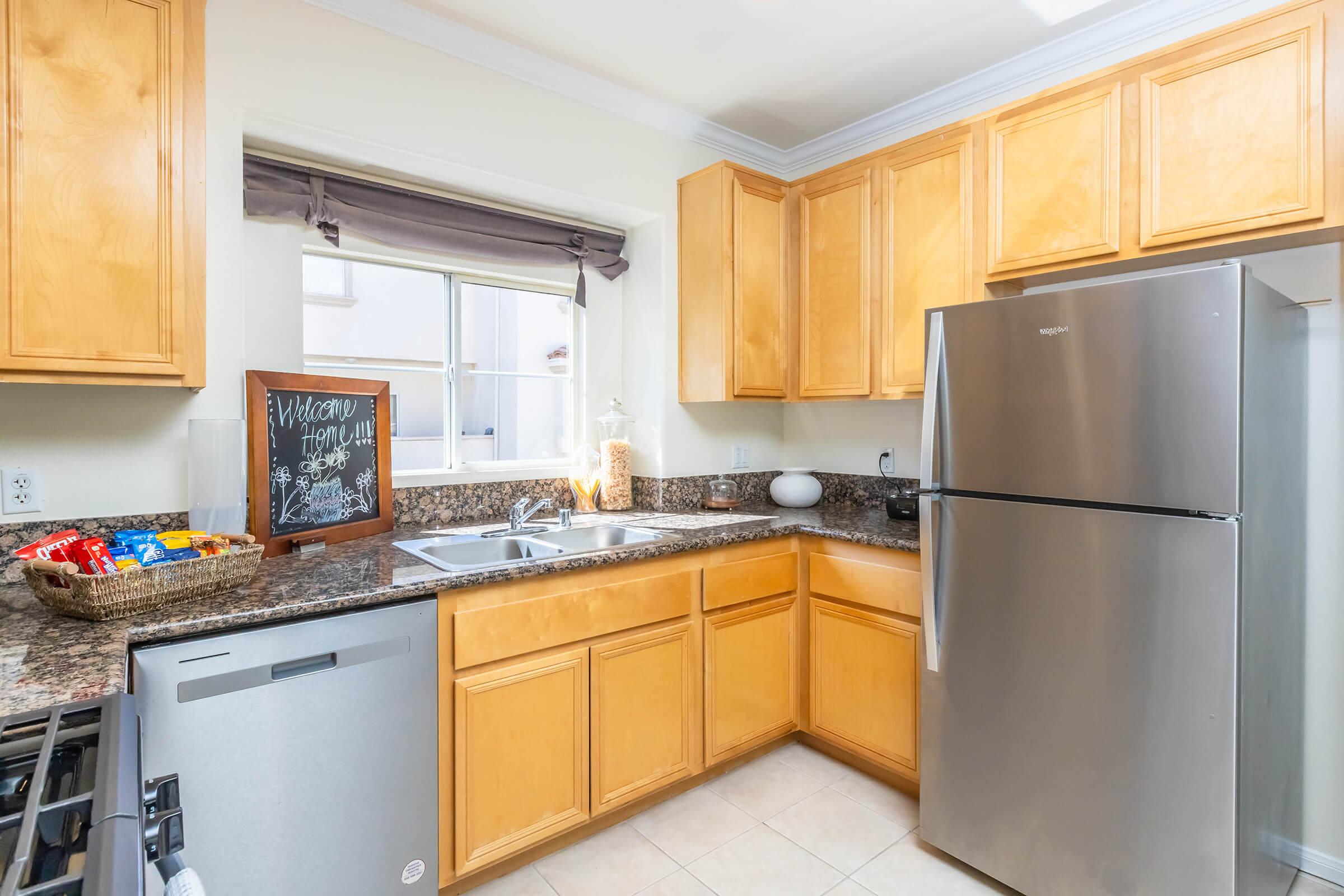 A modern kitchen featuring wooden cabinets, a stainless steel refrigerator, a double sink, and a granite countertop. There’s a chalkboard on the wall and a basket with snacks on the countertop. Natural light streams in through a nearby window, enhancing the warm, inviting atmosphere.