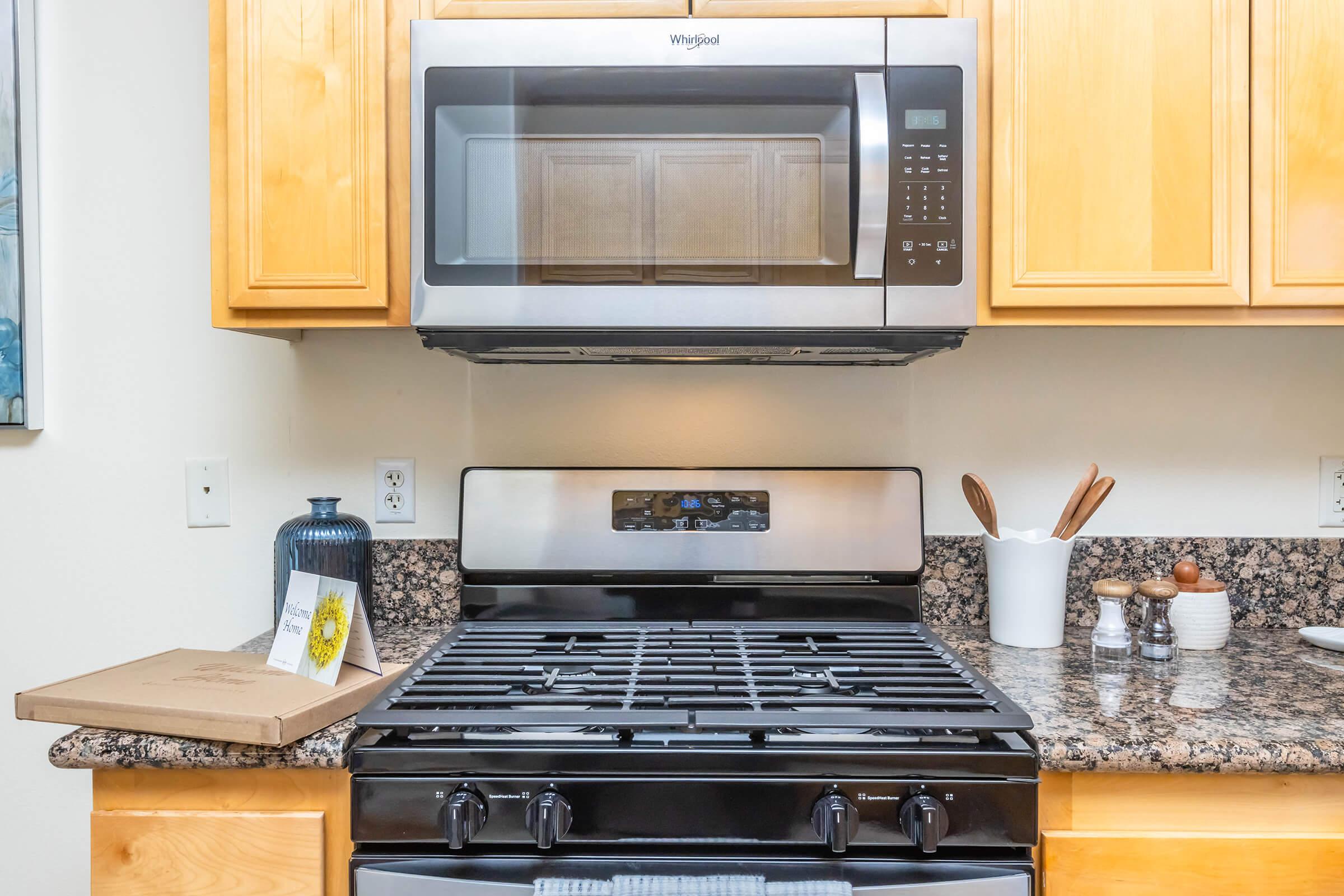 A modern kitchen featuring a stainless steel Whirlpool microwave mounted above a gas stove. The stove has a black griddle and knobs, with a countertop made of dark granite. Nearby, there are utensils in a holder and a decorative blue vase, along with a pizza box partially opened on the counter.