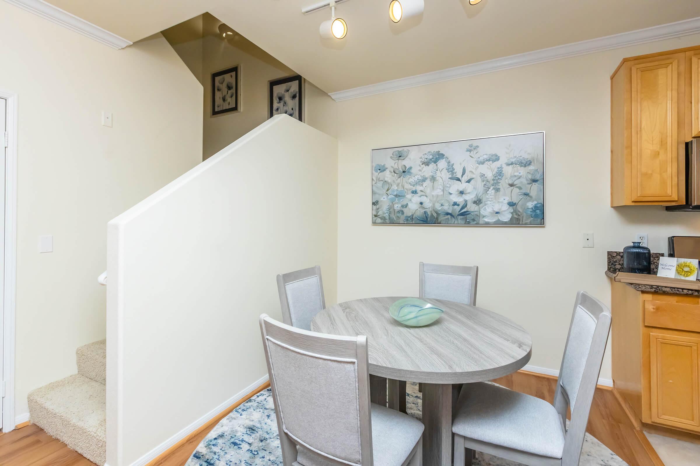 A cozy dining area with a round wooden table surrounded by four light gray chairs. A decorative bowl is placed in the center of the table. The background features a staircase leading to an upper level, with soft lighting and a floral artwork on the wall.