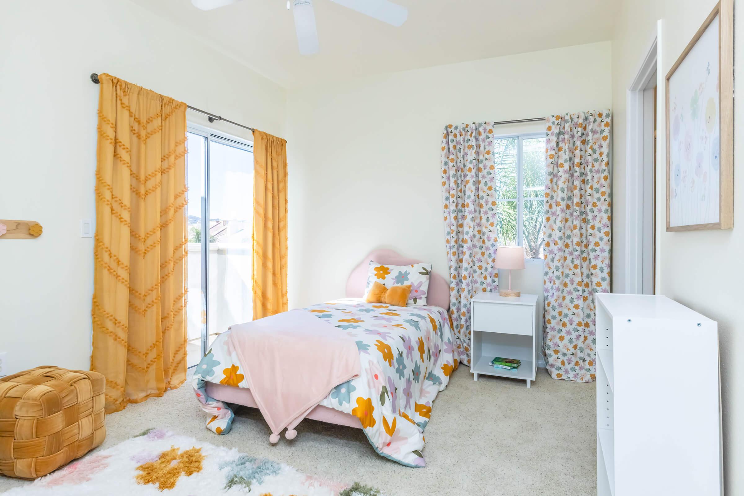 A bright and airy bedroom featuring a twin bed with a floral-patterned comforter, a small nightstand with a lamp, and light-colored curtains. The room includes a large window and a cozy rug, creating a warm and inviting atmosphere.