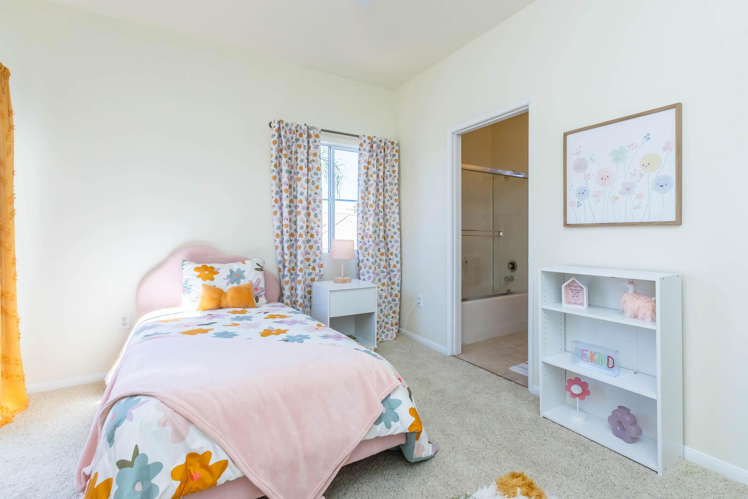A bright and cheerful bedroom featuring a bed with colorful floral bedding, a bedside table, and a window adorned with light curtains. There is a bathroom door visible, and a white bookshelf with decorative items. The room has soft carpet and a warm, inviting atmosphere.