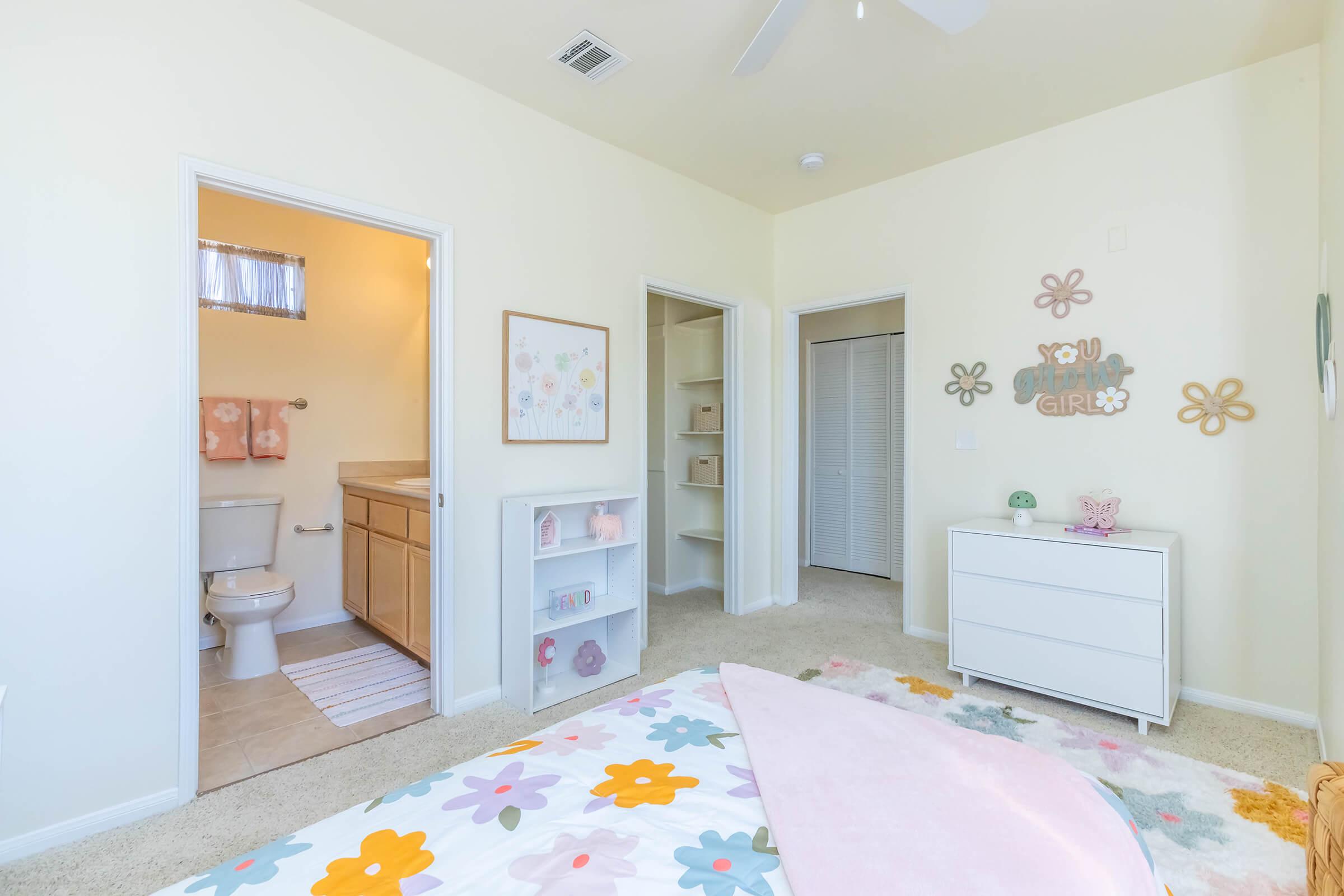 A bright and cheerful bedroom featuring a bed with colorful floral bedding, a dresser, and a small bookshelf. The walls are painted pale yellow, and there is a bathroom door on the left. The decor includes playful flower motifs and a sign that says "You Go Girl." Natural light fills the room through a nearby window.