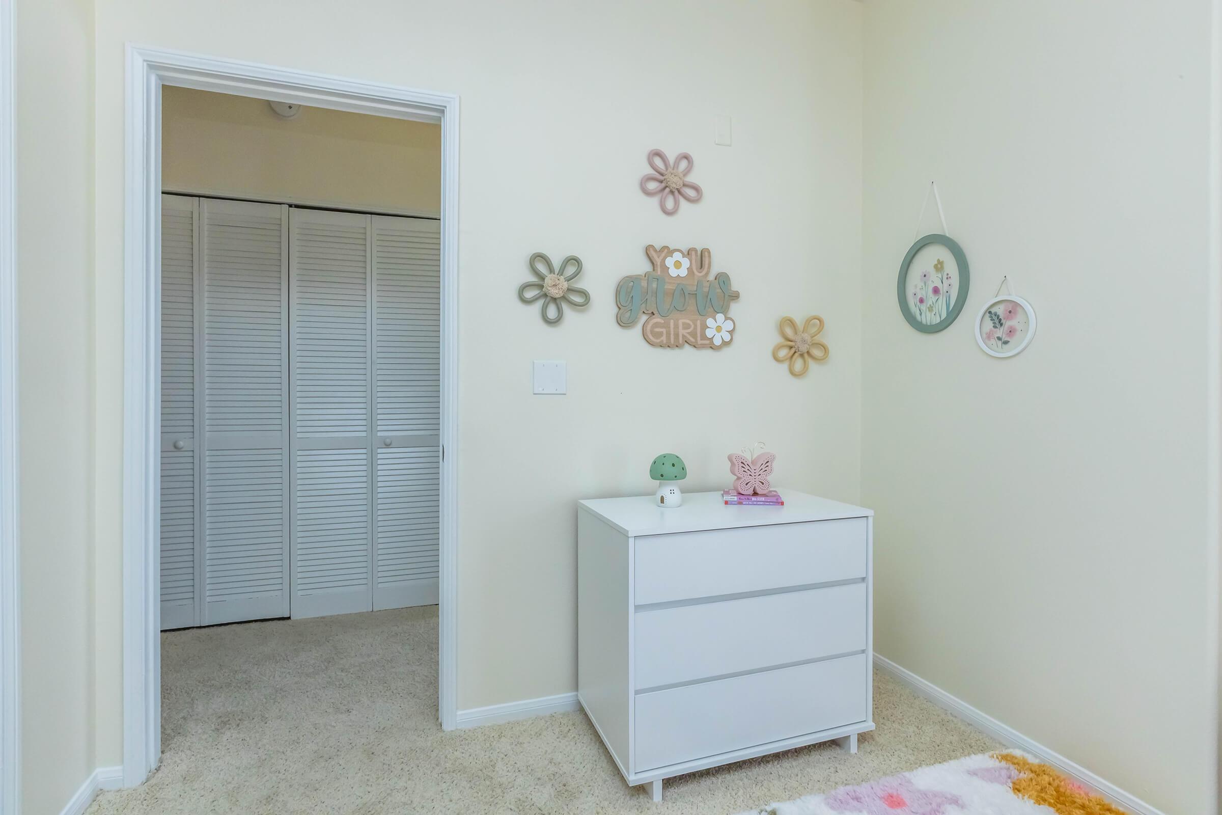 A cozy and bright children's room corner featuring a white dresser with a small decorative green mushroom and a butterfly. On the wall, colorful flower-shaped wall art and framed pictures convey a playful atmosphere. The room has light-colored walls and a carpeted floor, with a closet door visible.