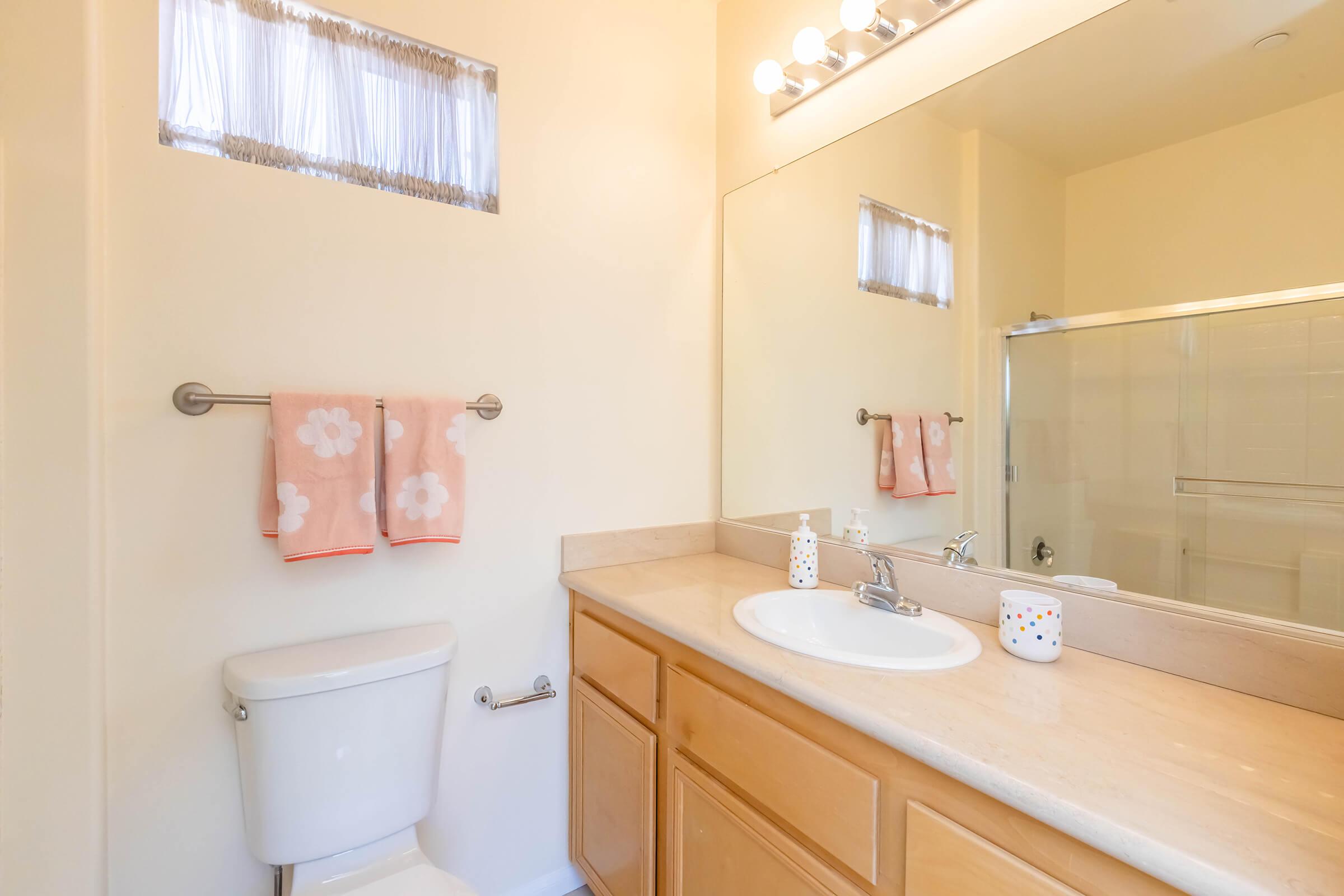 A bright and clean bathroom featuring a beige color scheme. The vanity has a circular sink and a large mirror above it. A shower enclosure is visible in the corner, and two light pink towels hang on a towel rack. The space is well-lit with natural light coming from a small window.