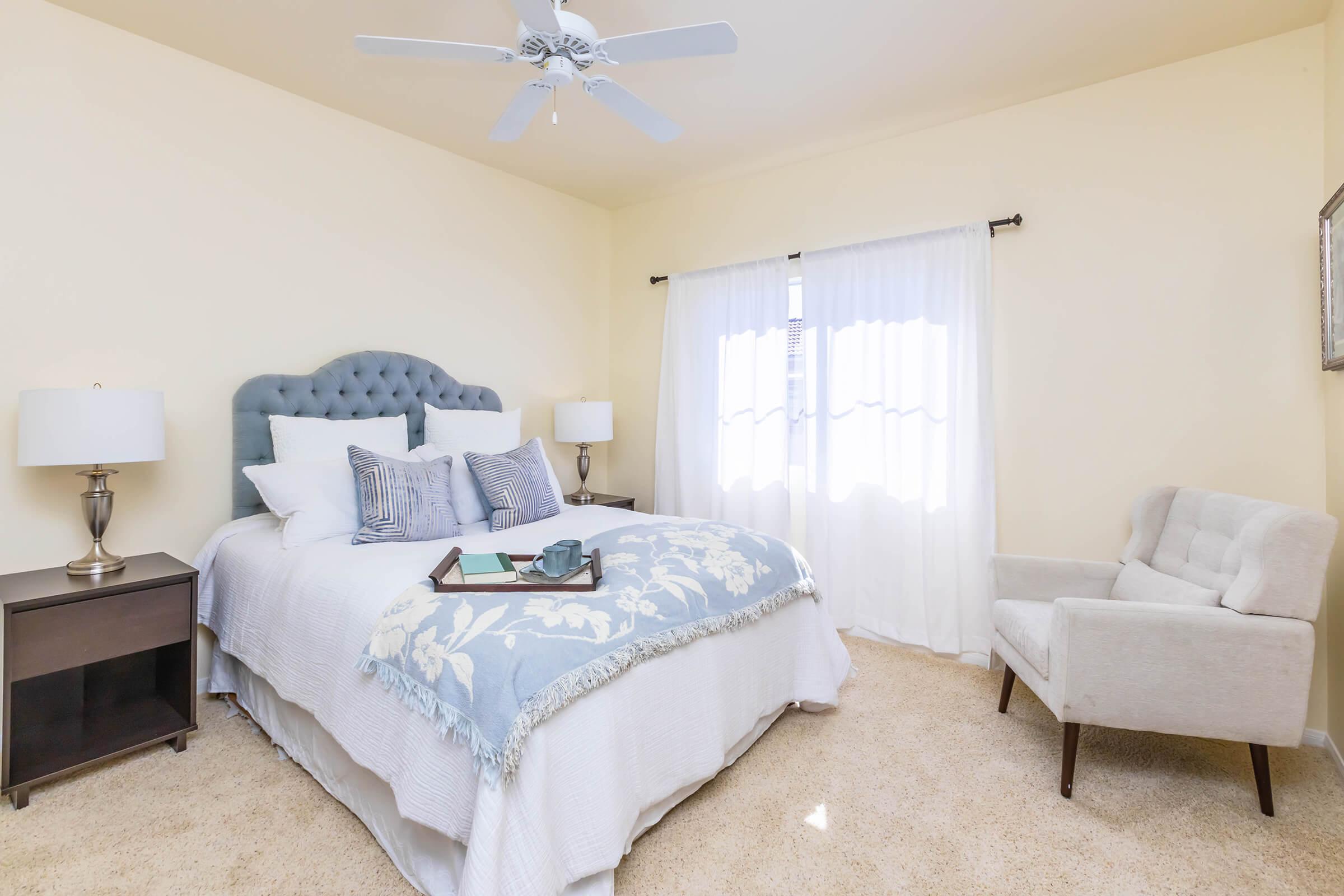 A cozy bedroom featuring a light-colored wall, a queen-sized bed with a blue and white comforter, accent pillows, and a tufted headboard. There's a nightstand with a lamp on one side and a comfortable armchair beside the window dressed with sheer curtains, creating a warm and inviting atmosphere.