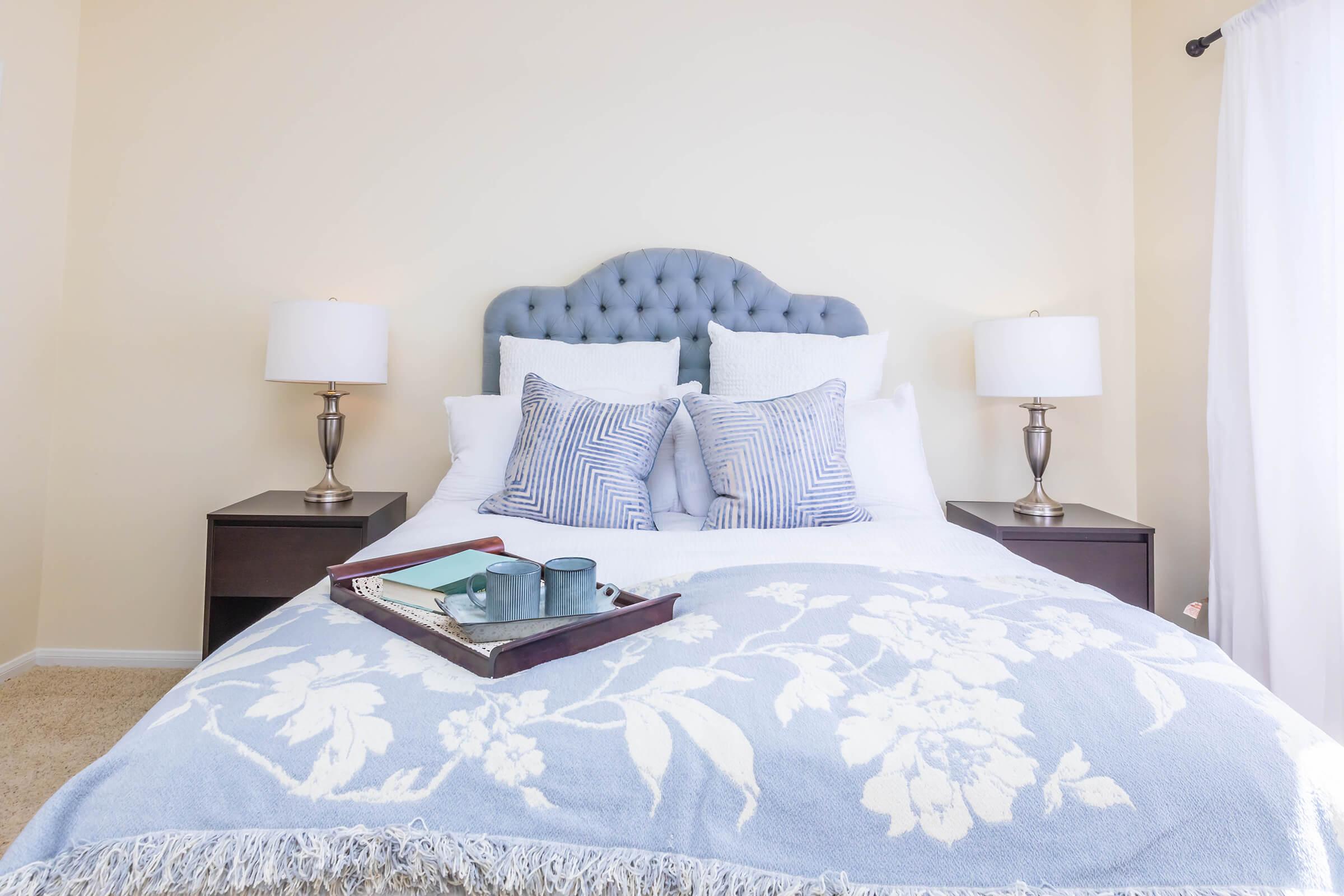 A well-decorated bedroom featuring a tufted blue headboard, white bedding with decorative pillows, and two bedside lamps. A tray with a book and decorative items sits on the bed, and the walls are a soft beige shade, creating a cozy atmosphere.