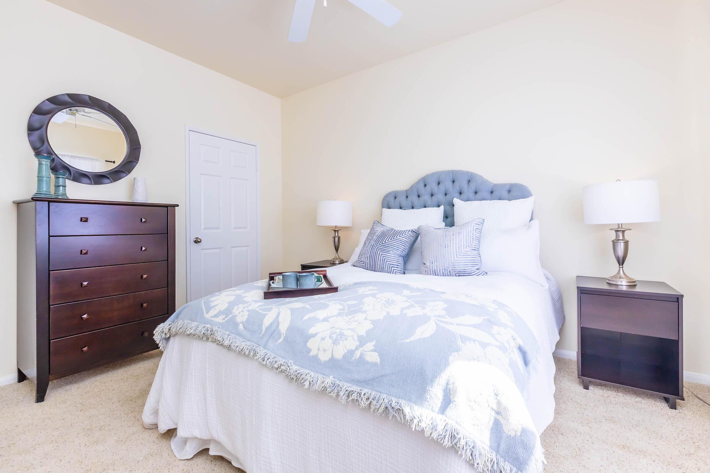 A cozy bedroom featuring a queen-sized bed with a light blue tufted headboard and white bedding, accented with a floral throw blanket. There are two bedside tables with lamps, a dark wooden dresser, and a round mirror on the wall, all set against soft beige walls and carpet.