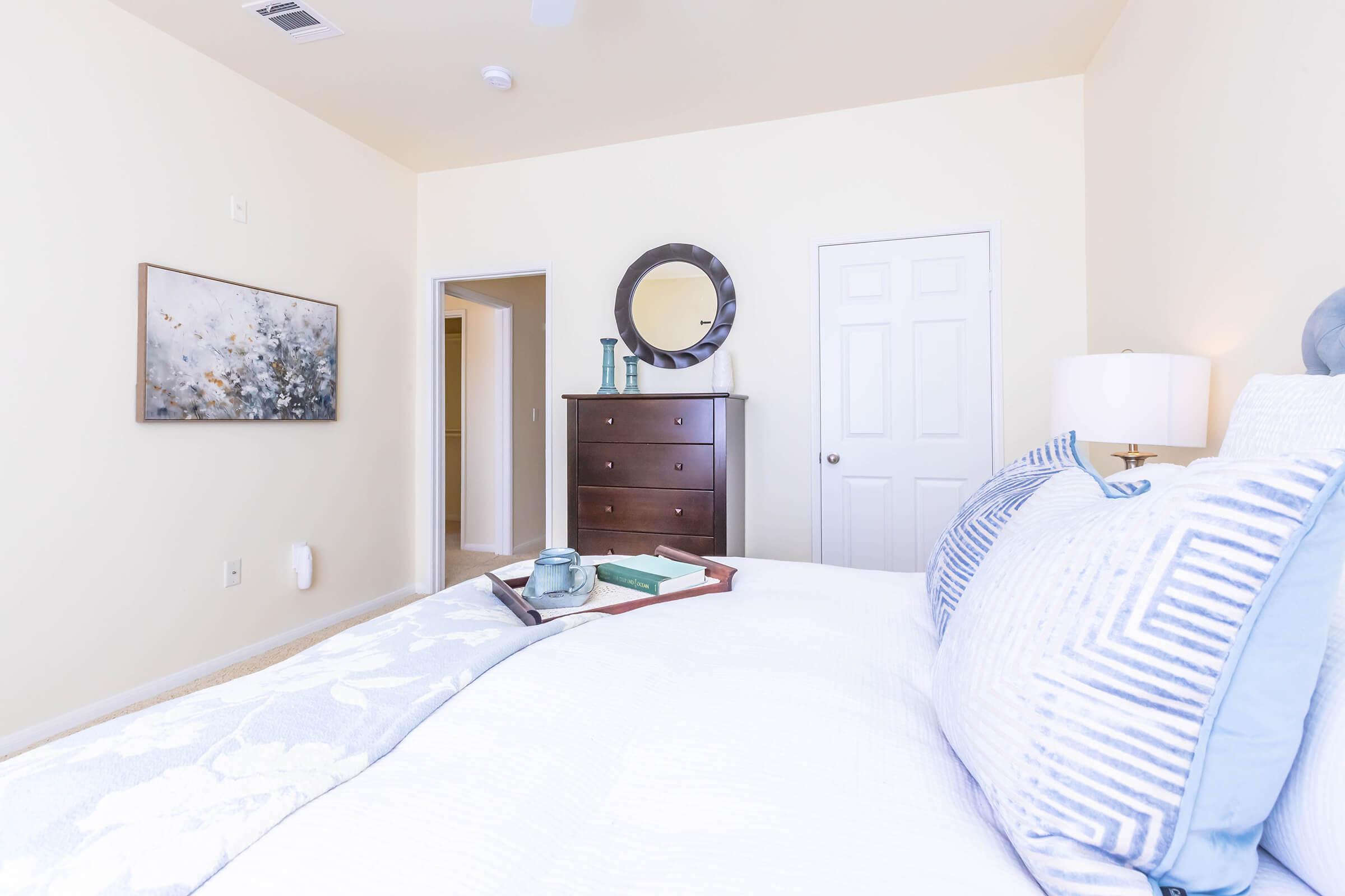 A cozy, well-lit bedroom featuring a neatly made bed with decorative pillows. A bedside table holds a tray with a cup and books. A dresser with a circular mirror is visible, and a piece of abstract art adorns the wall. The room has light-colored walls and warm, inviting decor.