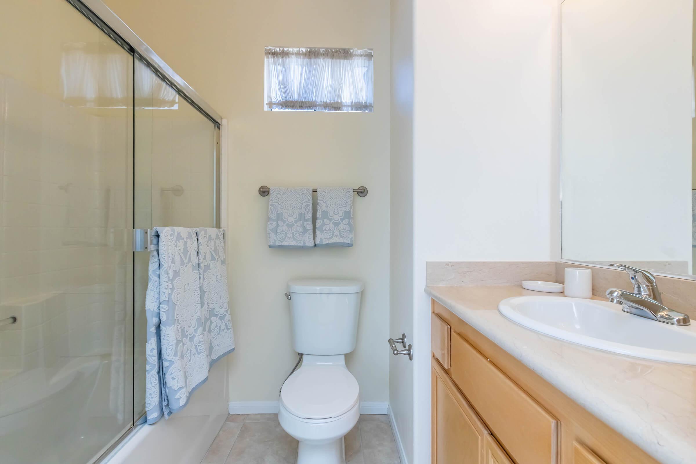 A clean and bright bathroom featuring a shower with a glass door, a toilet, and a wooden vanity with a sink. Two blue and white towels hang on a towel rack, and there is a small window providing natural light. The walls are painted a light color, adding to the spacious feel.