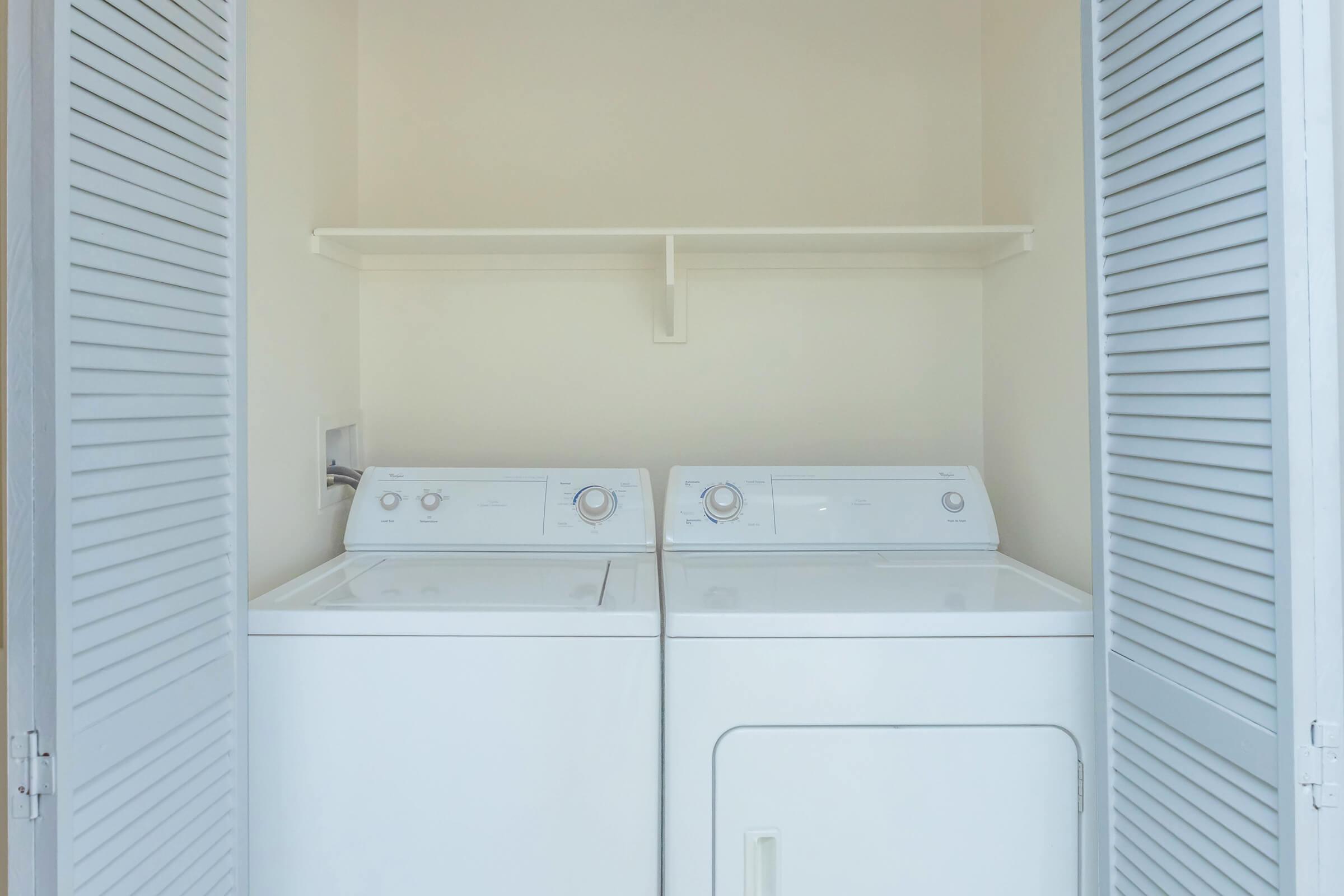 A clean laundry area featuring a white washing machine and dryer side by side, with control dials visible on the front. The machines are housed in a light-colored room with a shelf above them and closed louvered doors on either side.