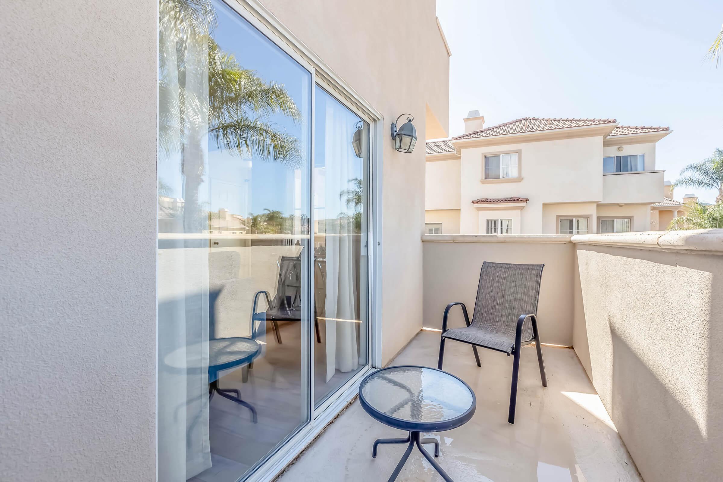 A small balcony featuring a glass table and two chairs, with sliding glass doors leading inside. The view includes a neighboring building and palm trees in the background, creating a relaxing outdoor space with ample natural light.