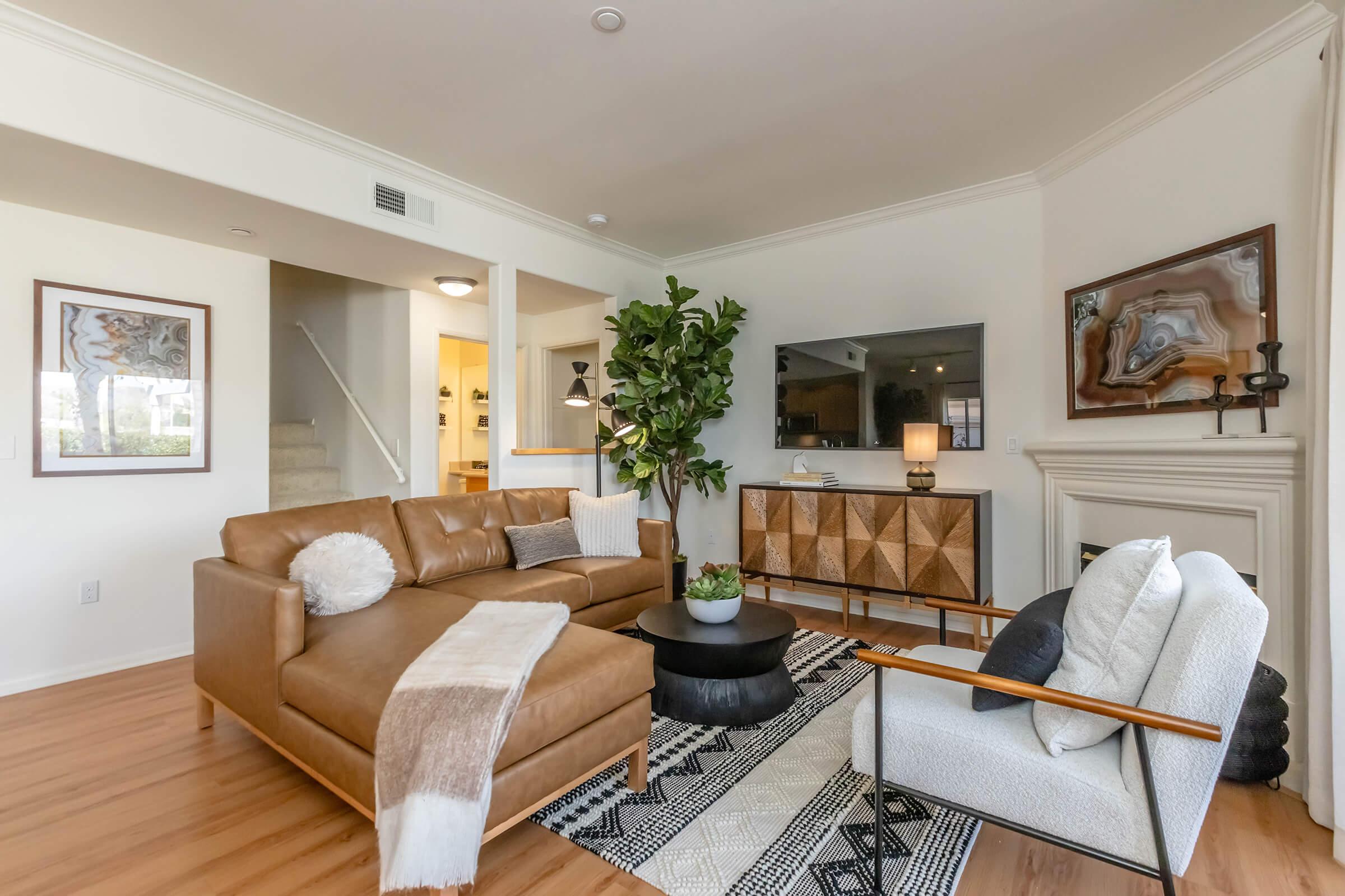 A modern living room featuring a brown leather sectional sofa with decorative cushions, a round black coffee table, and a stylish white armchair. A large indoor plant adds greenery, and a patterned rug enhances the decor. Wall art and a television are visible in the background, contributing to a cozy atmosphere.