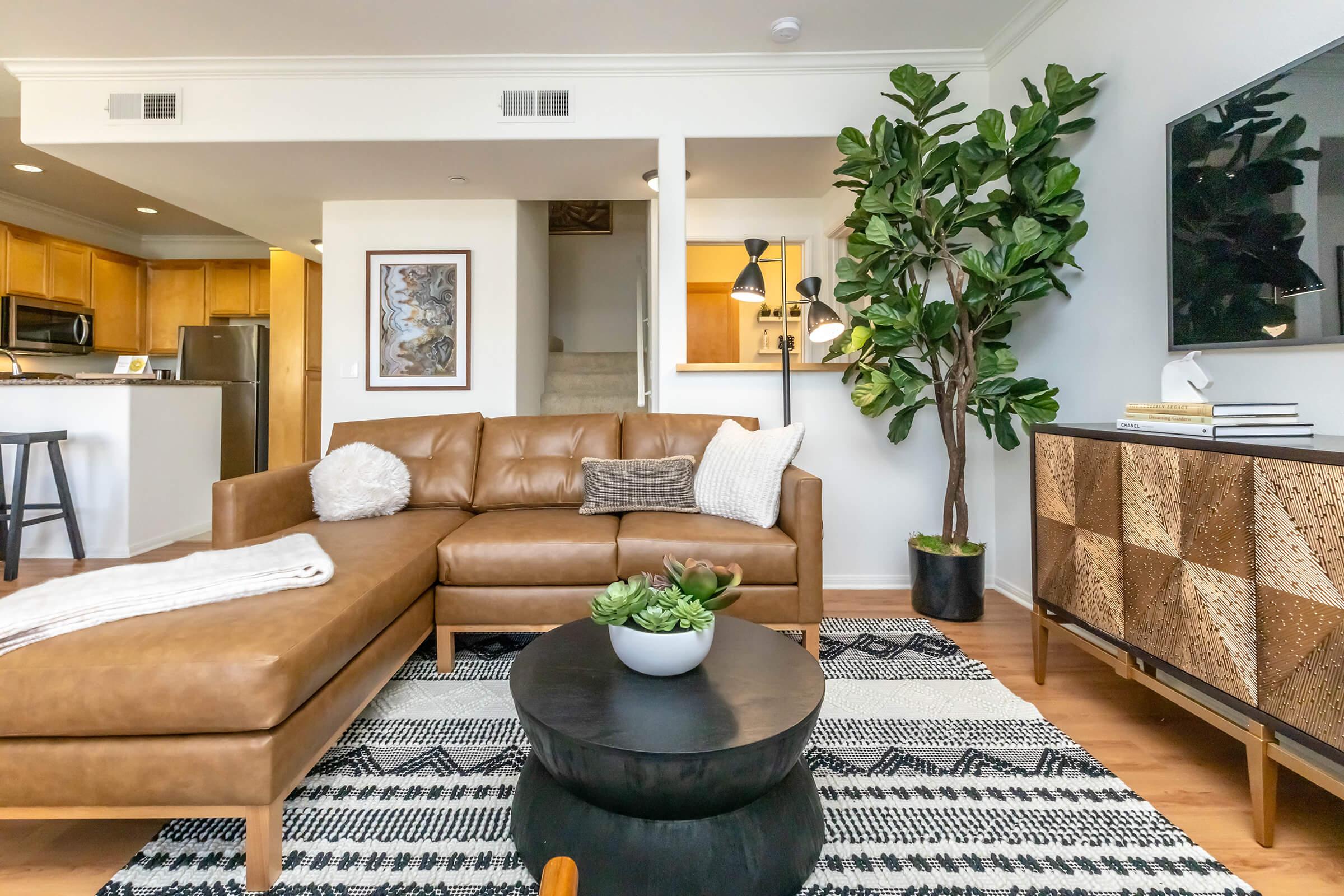A cozy living room featuring a brown leather sectional sofa with decorative pillows, an occasional chair, and a black coffee table. The space includes a large leafy plant, a patterned area rug, a sideboard with decorative elements, and a wall-mounted TV. The background shows a glimpse of a kitchen area.