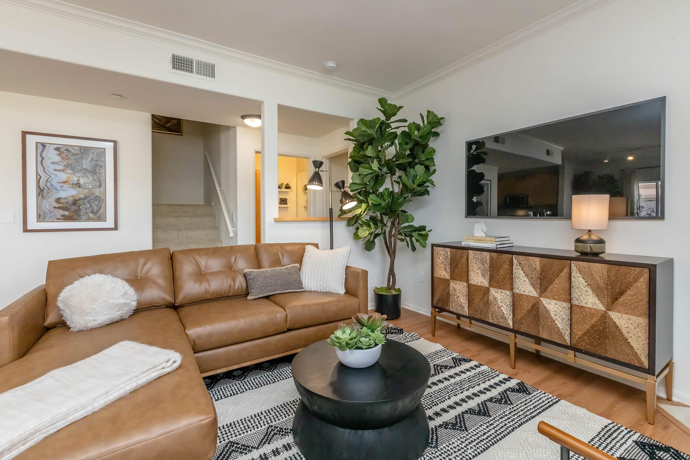 A cozy living room featuring a brown leather sectional sofa with plush cushions, a round black coffee table, and a decorative plant. A large TV is mounted on the wall above a stylish media console with a geometric pattern. The space is well-lit, with a warm and inviting atmosphere.
