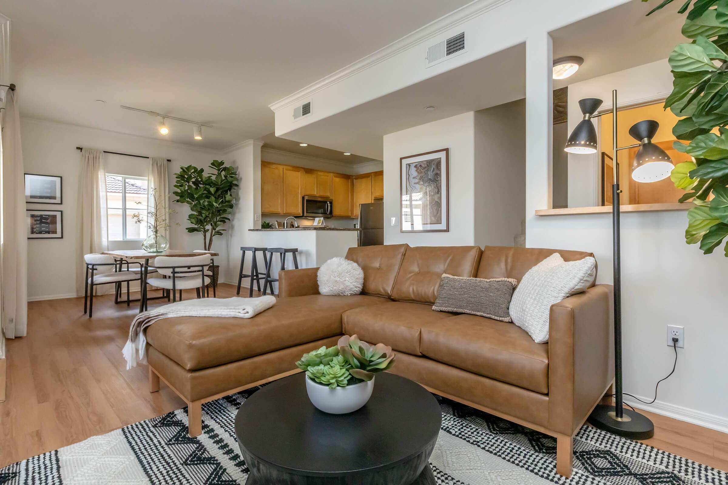 A modern living room featuring a brown leather sectional sofa adorned with decorative pillows, a round coffee table with a succulent plant, and a stylish rug. There is a dining area in the background with a small table and chairs, and the kitchen is visible with wooden cabinets and stainless steel appliances.