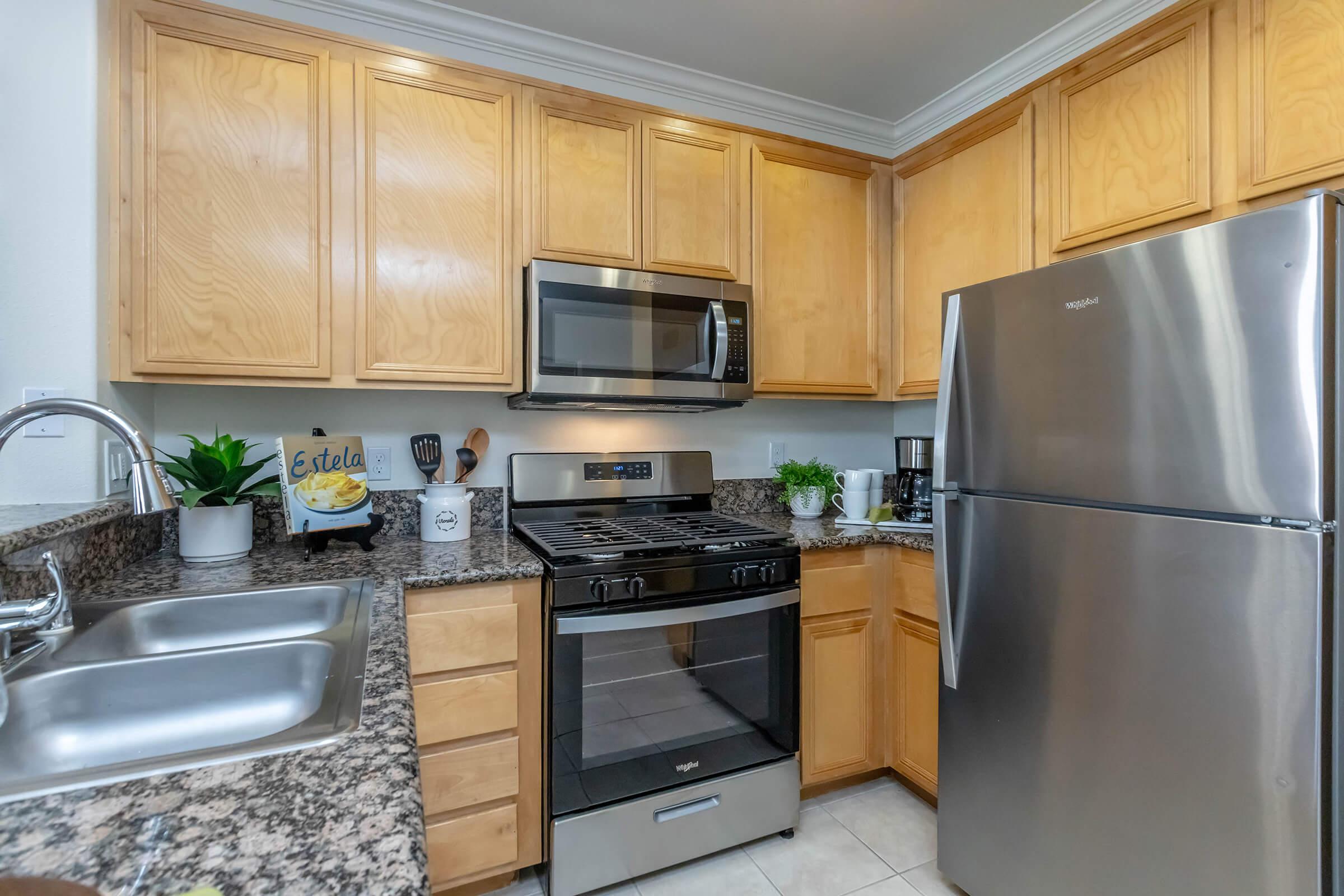 Modern kitchen featuring light wood cabinetry, a stainless steel refrigerator, a gas stove with an oven, and a microwave above. The countertops are granite, and there are decorative items and plants on display. Kitchenware and utensils are neatly arranged, emphasizing a clean and organized space.
