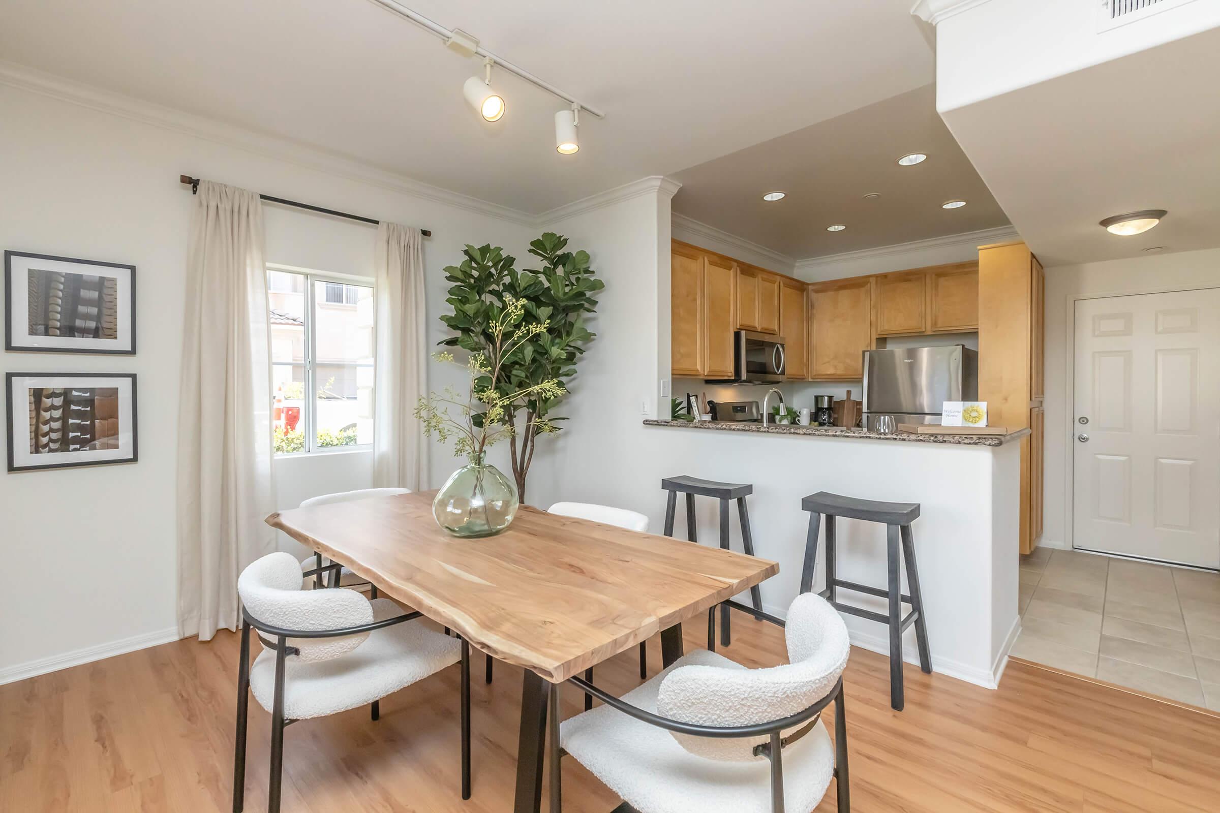 A modern dining area featuring a wooden table with four chairs, surrounded by light-colored walls and a large window with curtains. A small plant and a decorative vase are on the table. In the background, there's a kitchen with wooden cabinets and stainless steel appliances.