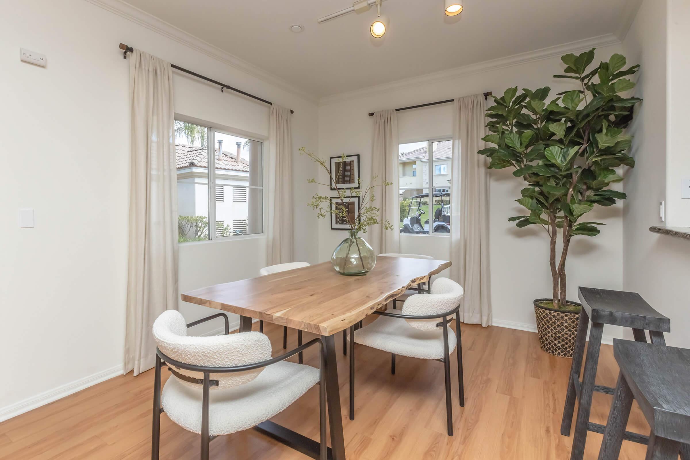 A bright dining area featuring a wooden table surrounded by four upholstered chairs. Large windows with light curtains allow natural light to fill the space. A potted plant adds greenery, and a minimalist side table is positioned against the wall. The walls are painted in a soft, neutral tone.