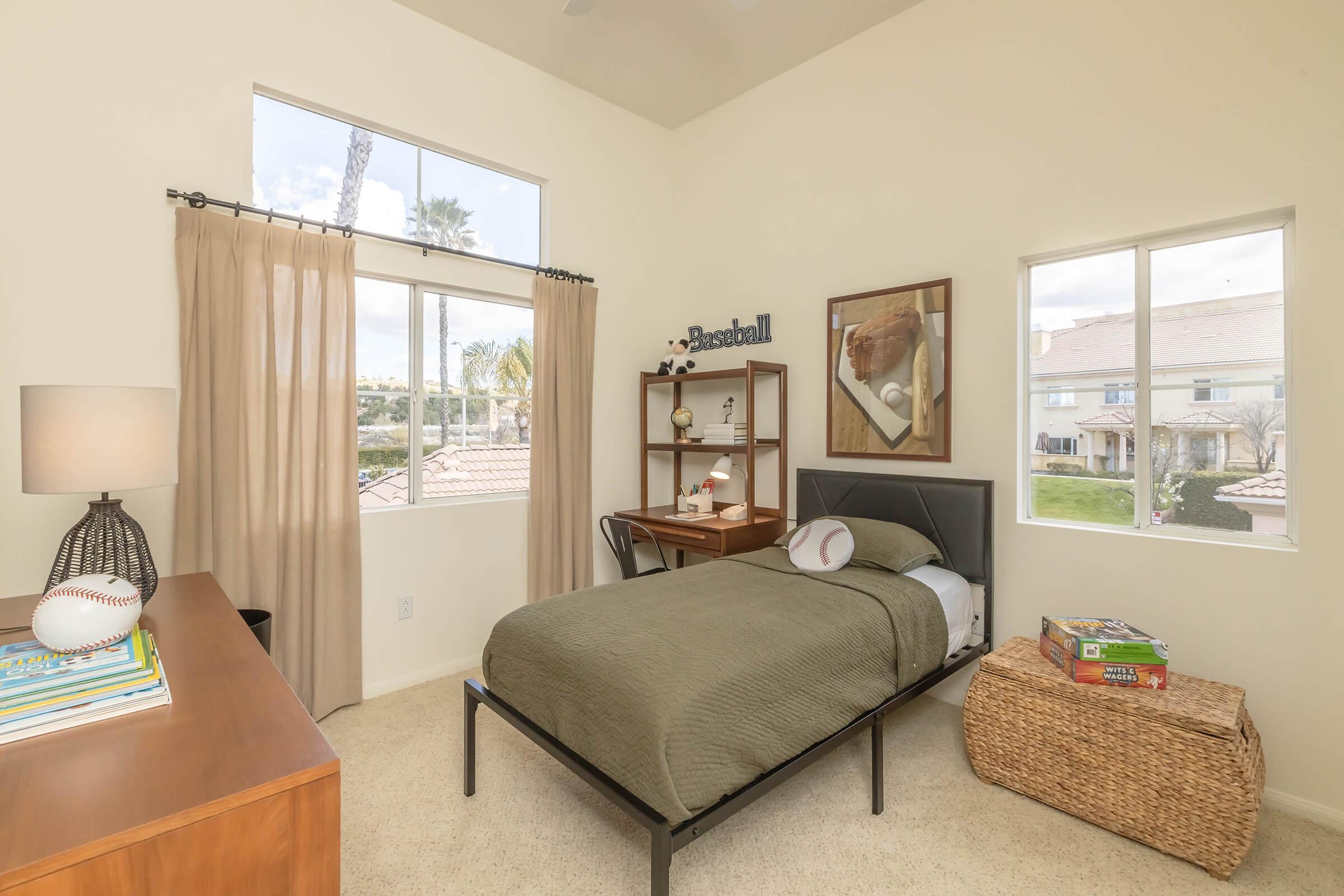 A cozy bedroom featuring a bed with green bedding, a nightstand with books, and a decorative baseball. The room has large windows allowing natural light, with palm trees visible outside. A bookshelf and a storage basket complete the simple and inviting decor.