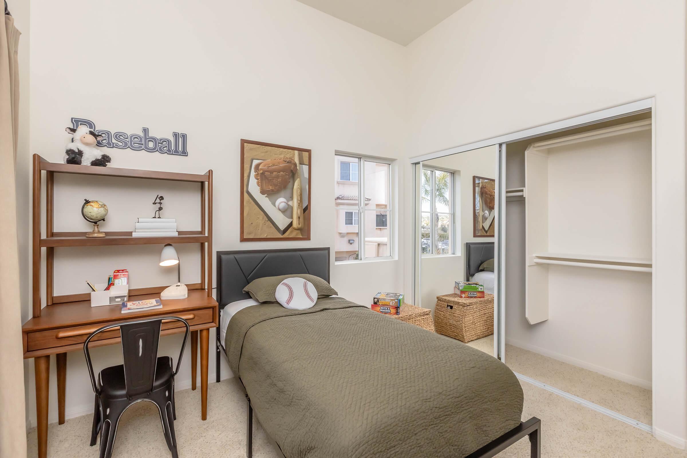 A cozy bedroom featuring a bed with a green quilt, a wooden desk with a globe and lamp, and baseball-themed artwork on the walls. A closet and a window allow natural light to fill the room, creating a bright, welcoming atmosphere. Baskets and books add a touch of decor and functionality.
