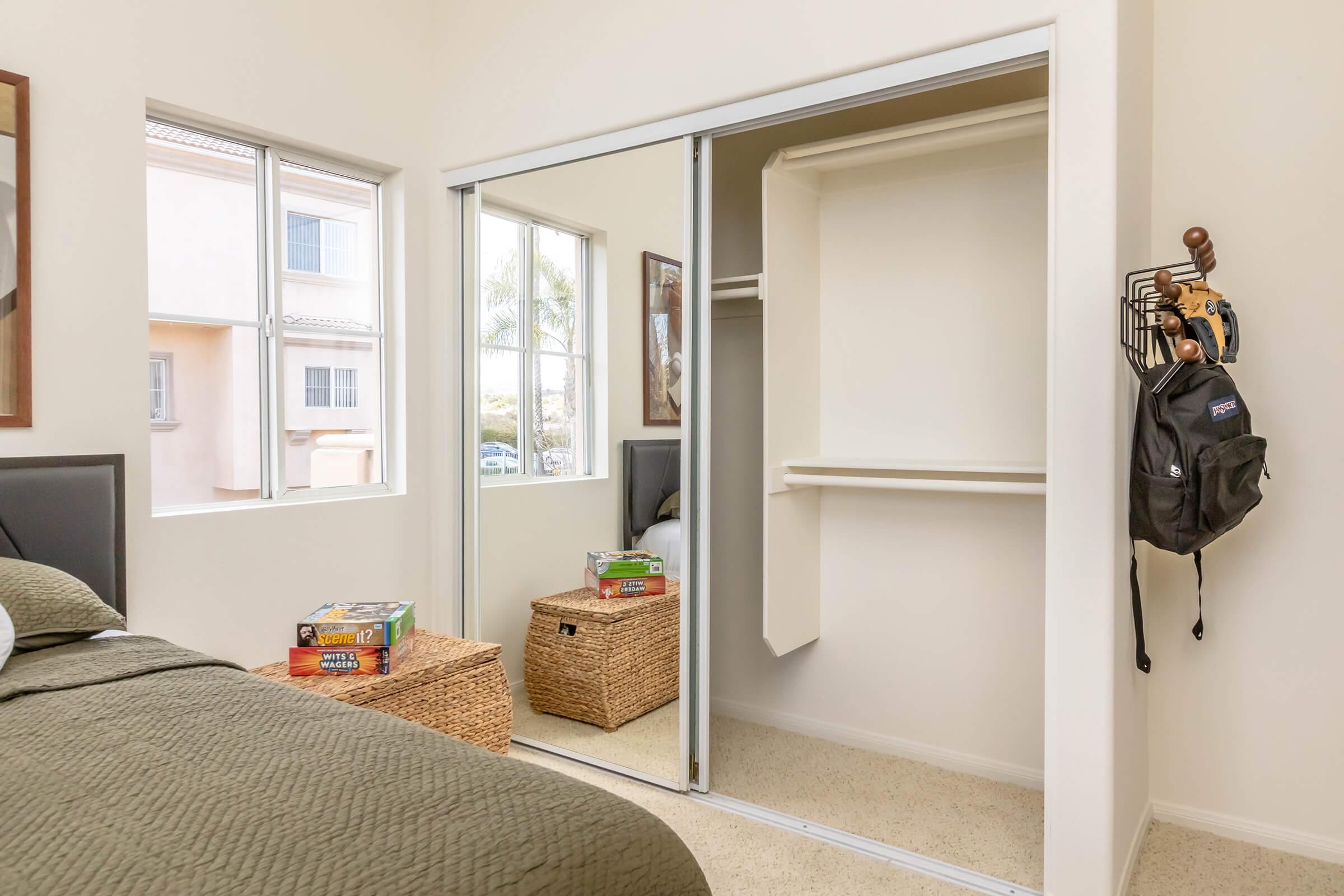 A cozy bedroom featuring a bed with green bedding, a mirror closet with sliding doors, and two woven baskets. Natural light enters through a window, illuminating the space. A backpack hangs on a hook beside the closet, while games are placed on top of the baskets. The overall ambiance is inviting and organized.