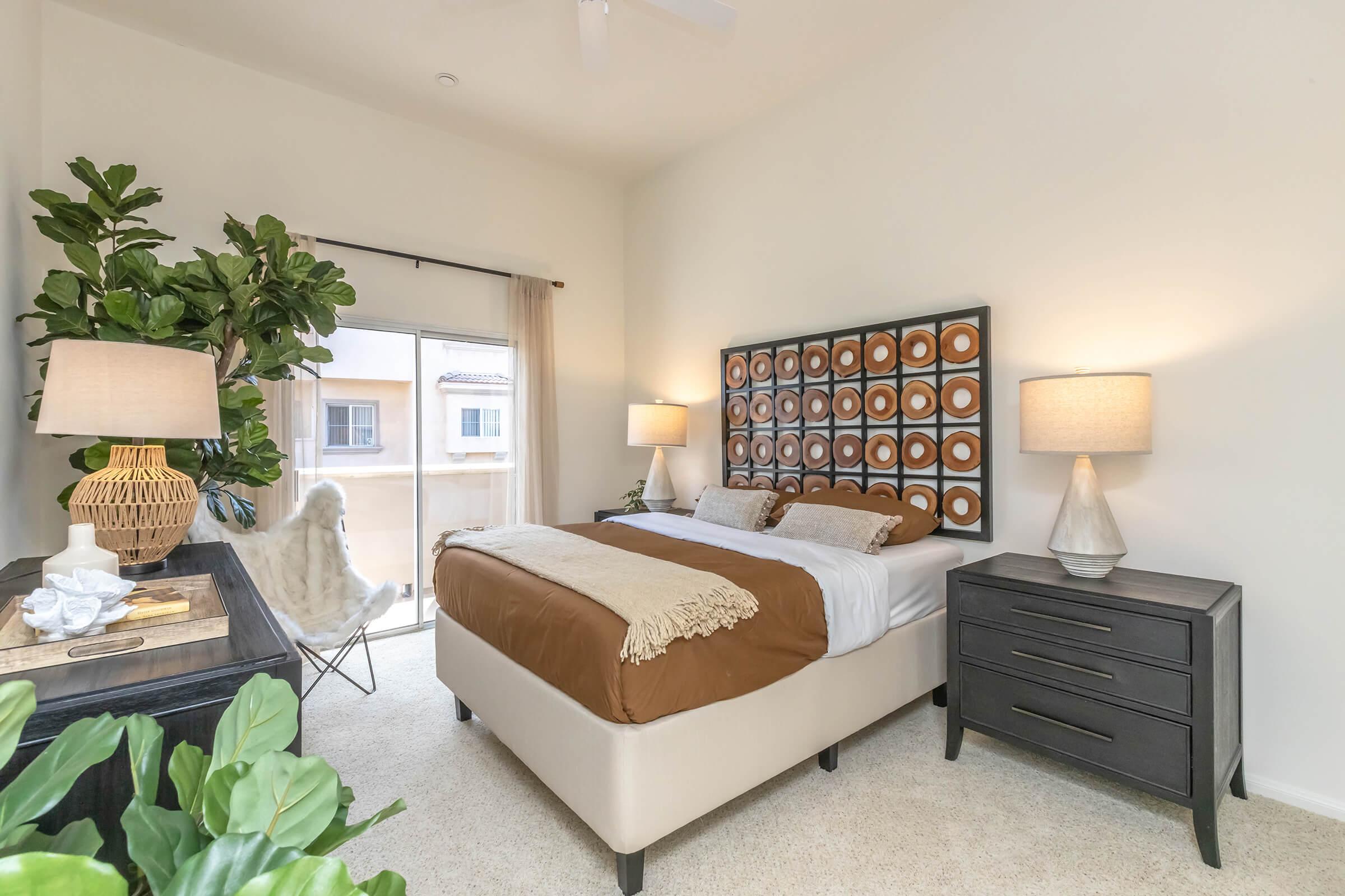 Cozy bedroom featuring a queen-sized bed with a brown and white bedding set, a decorative headboard, and two bedside lamps. There’s a stylish nightstand and a large window with sheer curtains. A plant adds a touch of greenery, enhancing the warm and inviting atmosphere of the space.
