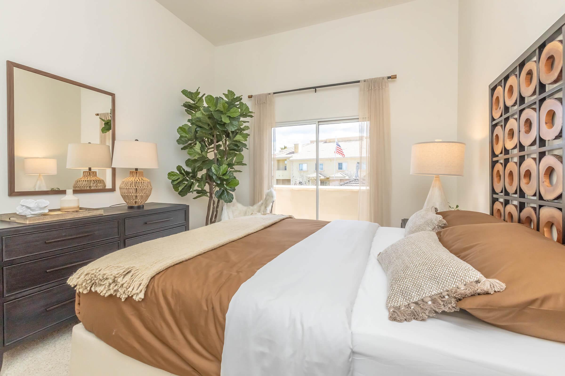 A beautifully styled bedroom featuring a king-sized bed with brown and white bedding, decorative pillows, bedside lamps, and a large mirror. A tall, leafy plant adds a touch of greenery, while sheer curtains frame a window with a view outside. A modern dresser completes the serene decor.