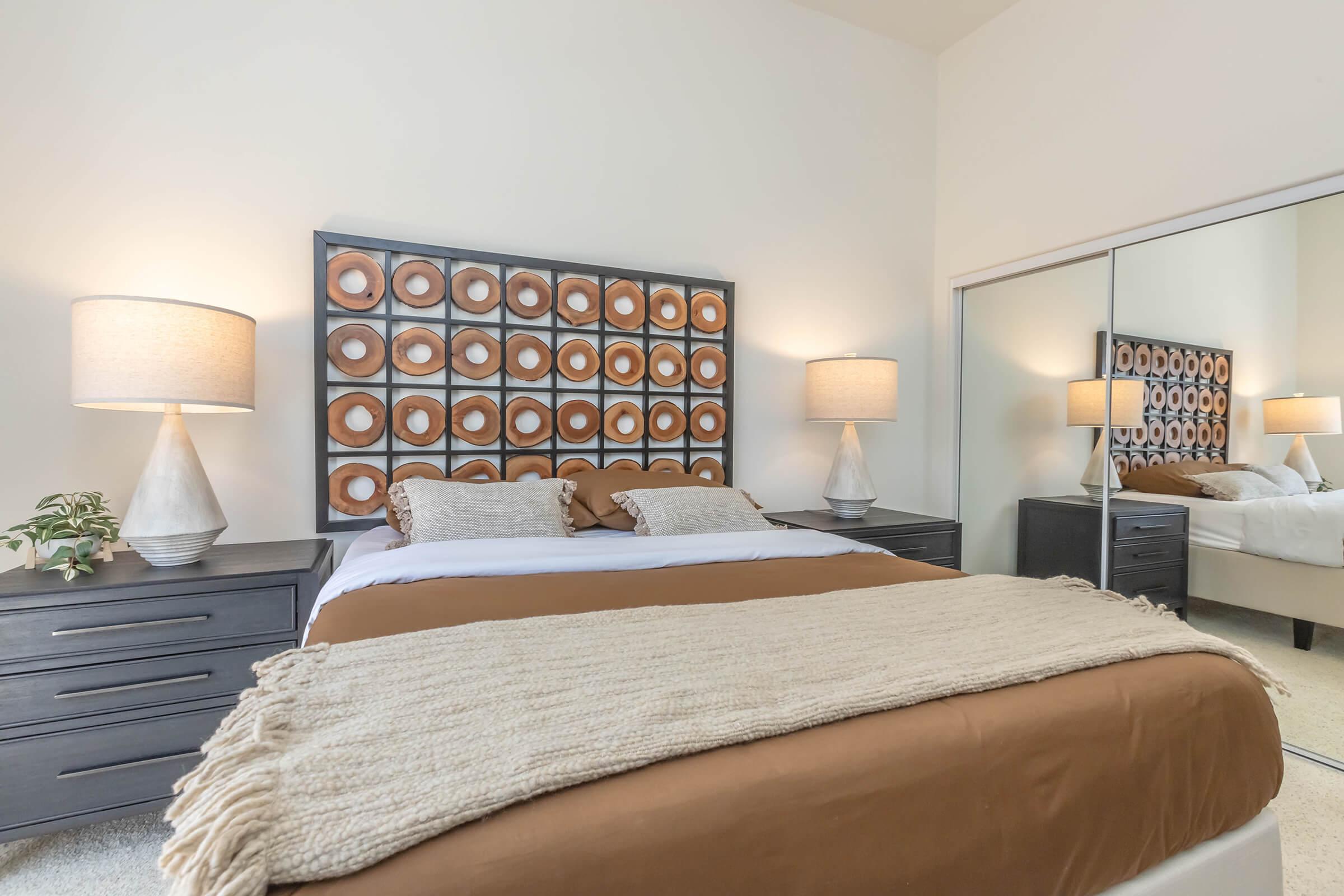 A cozy bedroom featuring a brown and white color scheme. The bed is adorned with decorative pillows and a textured blanket. On either side of the bed are two lamps with beige shades. A mirrored closet reflects the room's light, while a piece of abstract art hangs above the headboard, adding visual interest.