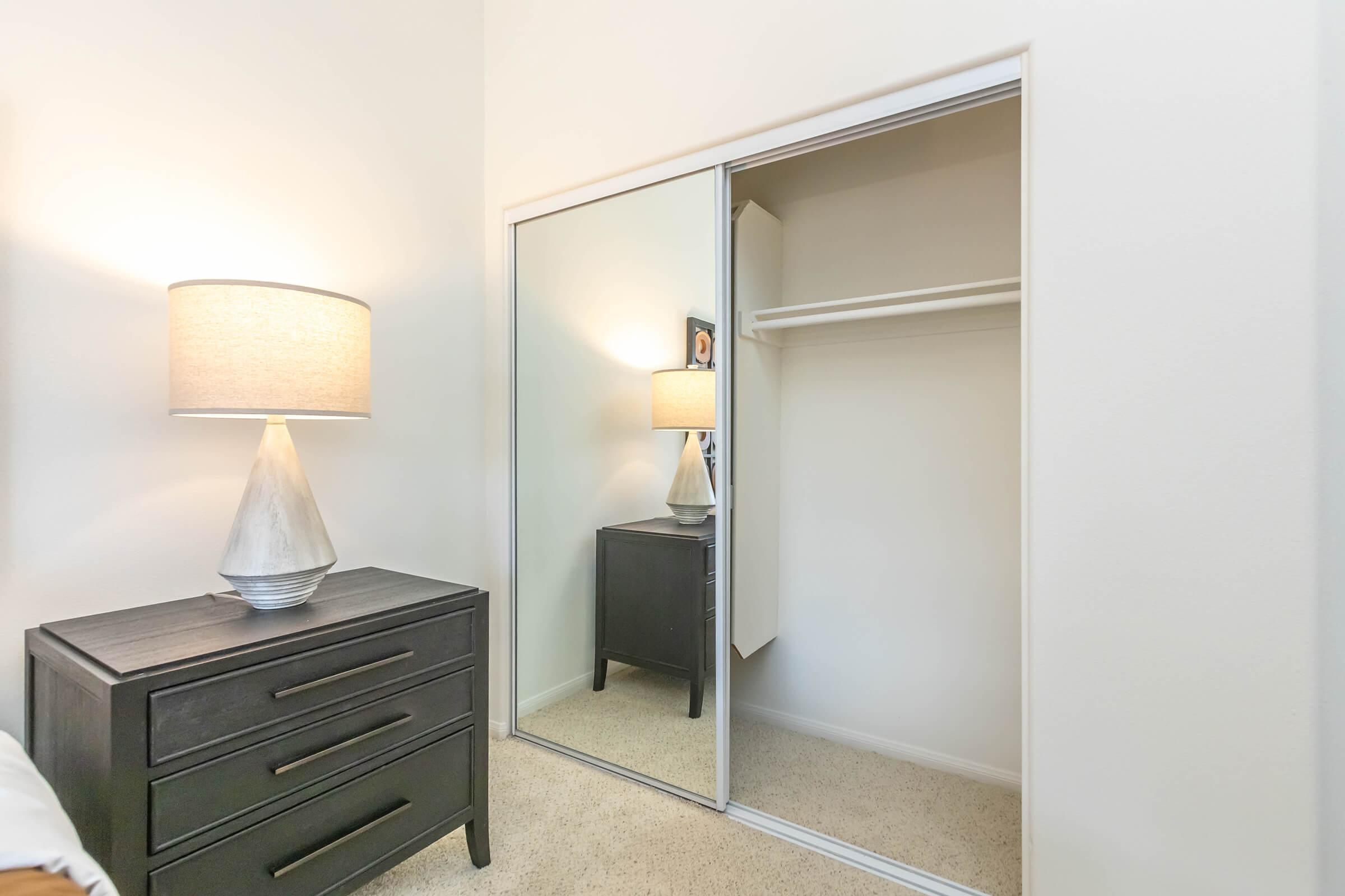 A well-lit room featuring a mirrored closet, a bedside table with a lamp, and neutral-colored walls. The floor is carpeted, and the decor is simple and modern, creating a cozy atmosphere. The closet is empty, showcasing its spaciousness.