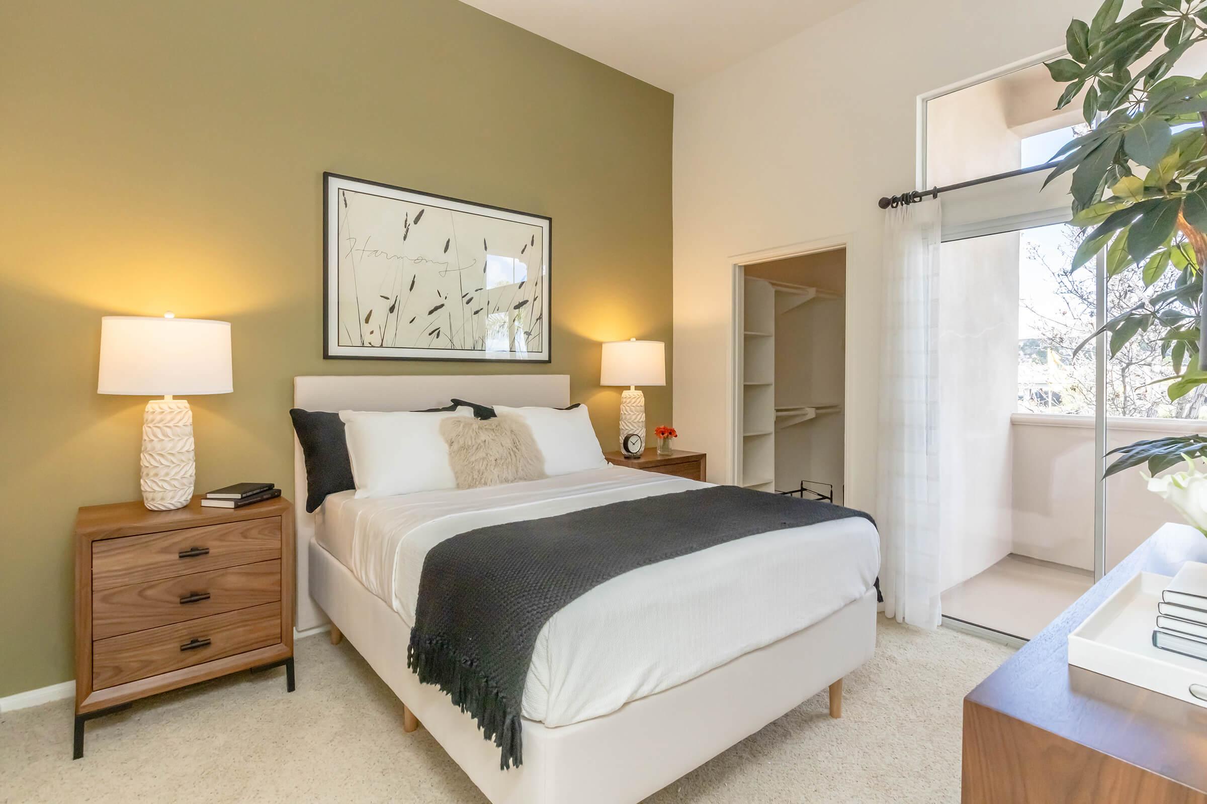A cozy bedroom featuring a queen-sized bed with a white duvet, accented by a dark throw blanket. Two bedside tables with lamps are on either side. A large window allows natural light, and there's a small balcony view. The walls are painted a soft green, creating a warm atmosphere.
