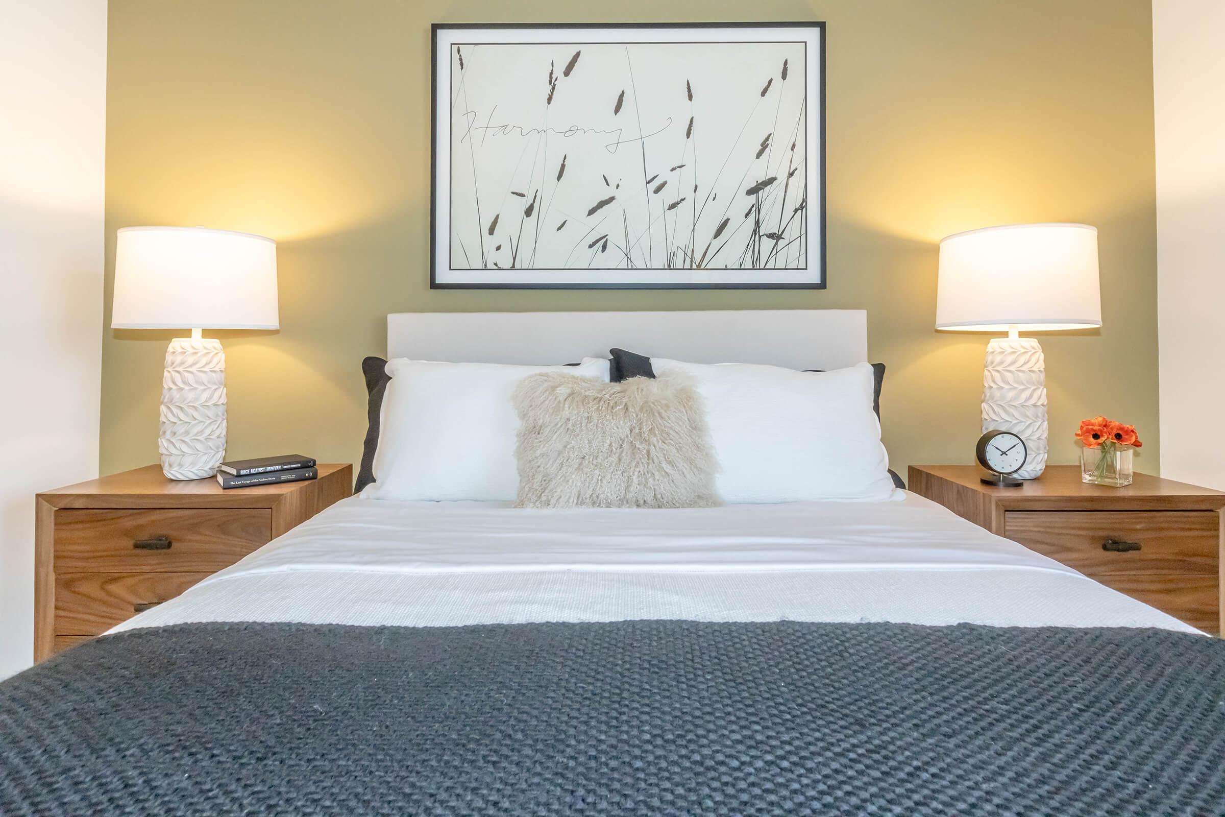 A cozy bedroom featuring a neatly made bed with white bedding and a textured blanket. On either side of the bed are wooden nightstands with decorative lamps. A framed artwork of grasses hangs above the bed, complemented by a clock and a small vase of flowers on one nightstand. Soft, warm lighting creates an inviting atmosphere.