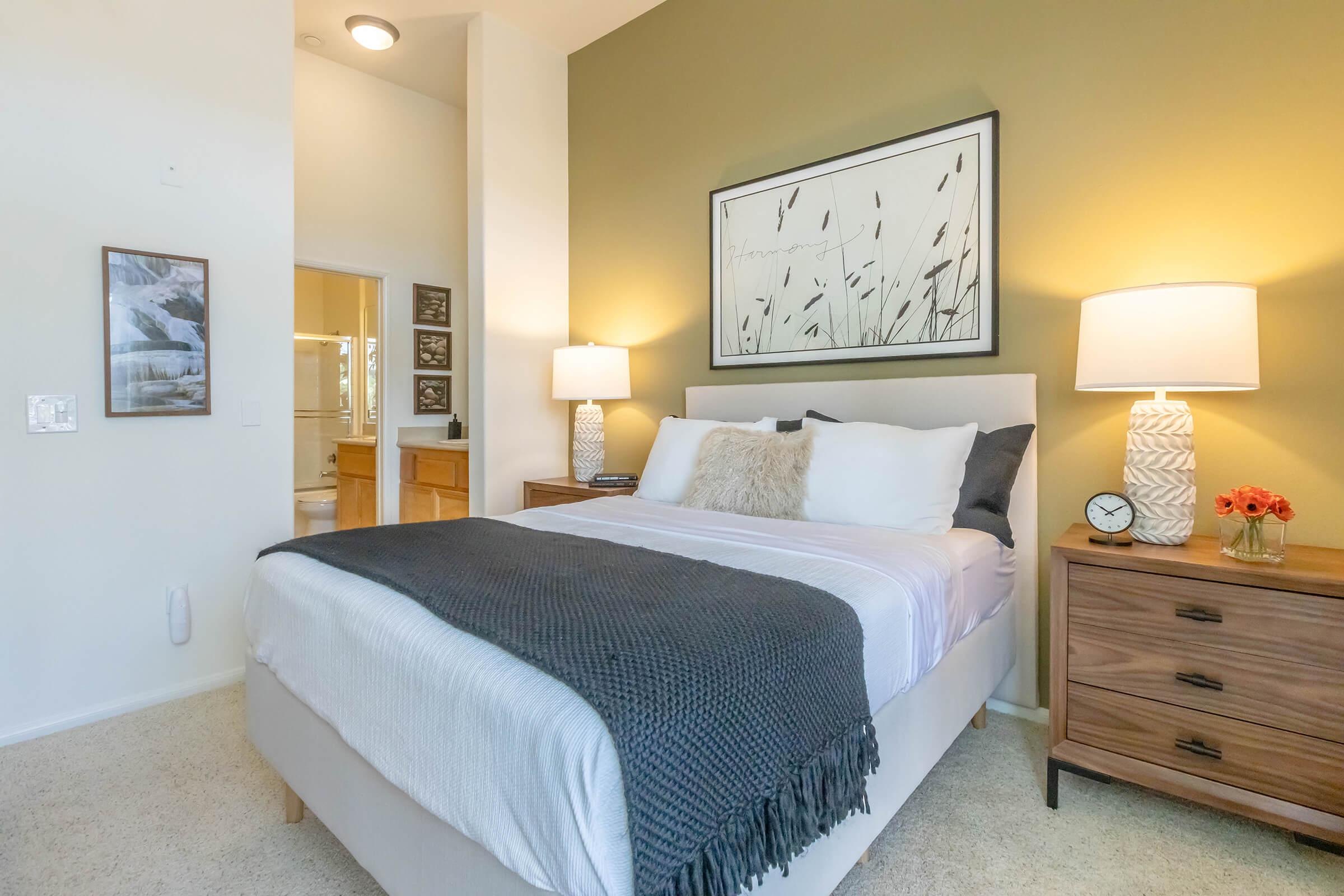 A cozy bedroom featuring a queen-sized bed with white linens and a dark throw blanket. There are two bedside tables with lamps, a decorative clock, and a small vase of flowers. The walls are painted a warm green, and a framed print of tall grass is hung above the bed. A doorway leads to a bathroom in the background.
