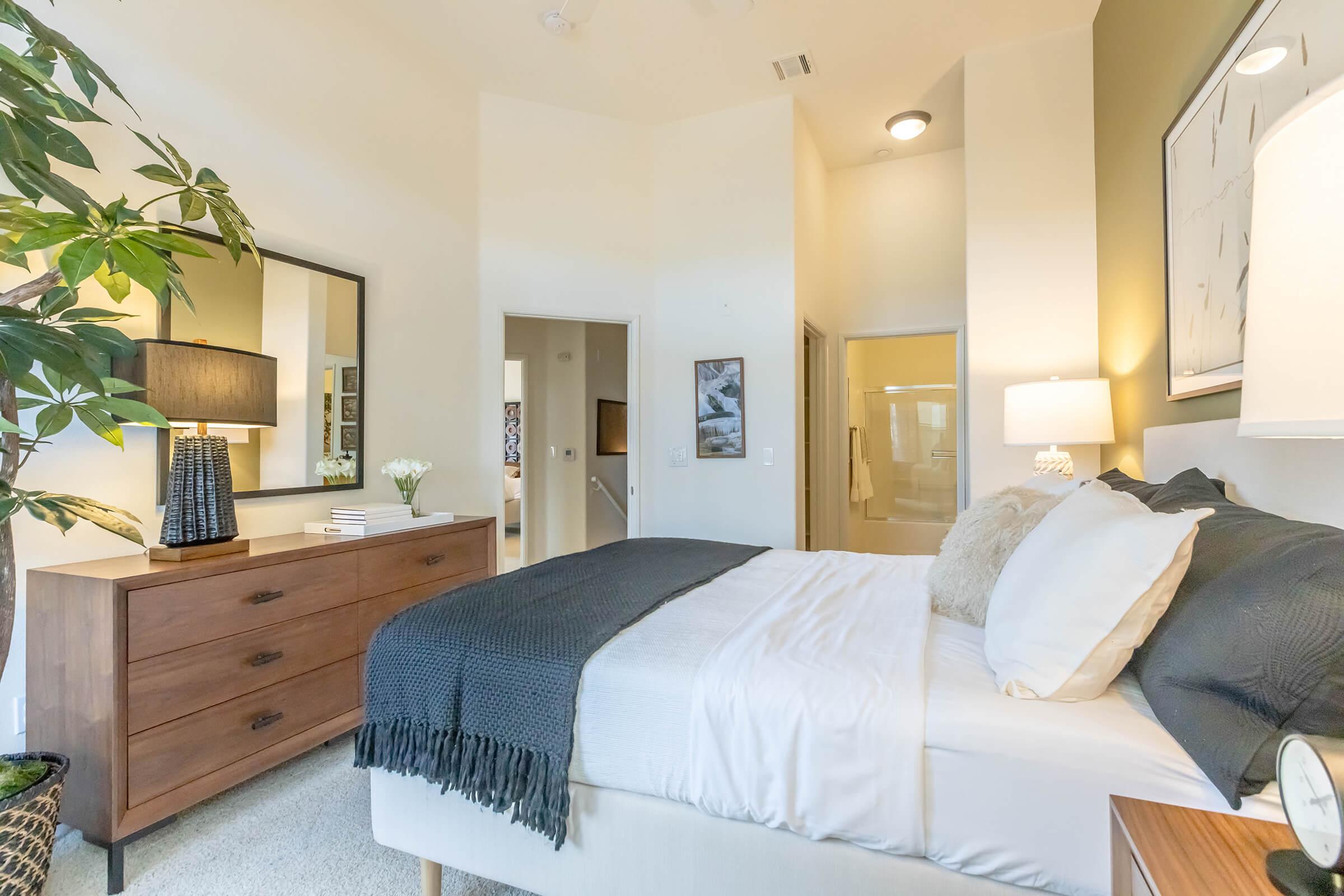 A cozy, well-lit bedroom featuring a large bed with white and gray bedding, a wooden dresser, and a stylish mirror. A lamp with a textured base sits on the nightstand, while a leafy plant adds a touch of greenery. The walls are a soft color, contributing to a tranquil atmosphere.