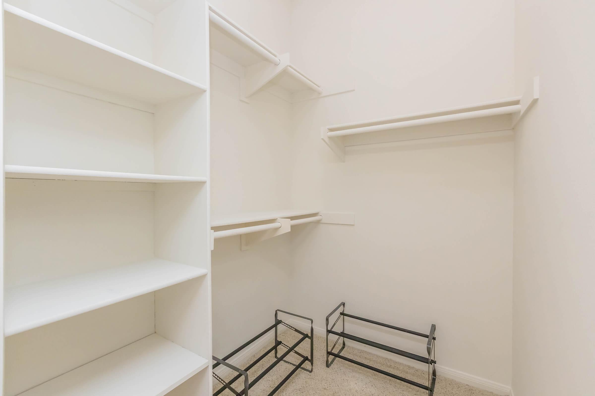 Empty closet with white shelves and a bare floor, featuring some black metal racks in the corner. The walls are painted light, and there is ample space for storage.