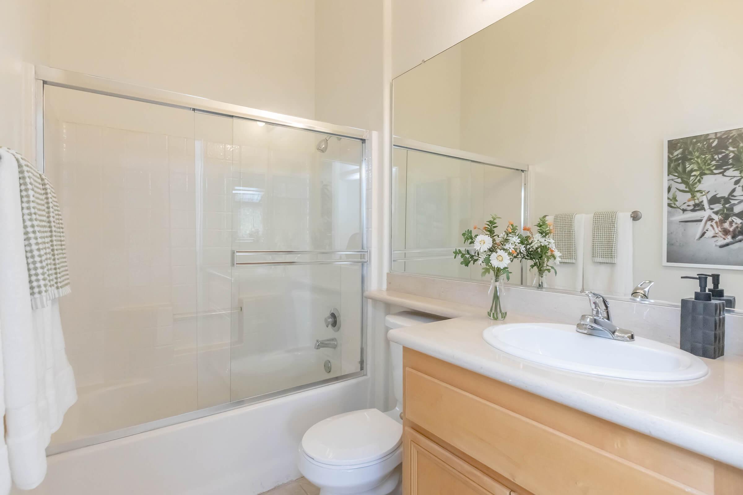 Bright and clean bathroom featuring a glass shower enclosure, a white sink with a silver faucet, and wooden cabinetry. A vase with flowers adds a touch of color, and a small framed photo of greenery is on the wall. Fresh towels are neatly hung, enhancing the inviting atmosphere.