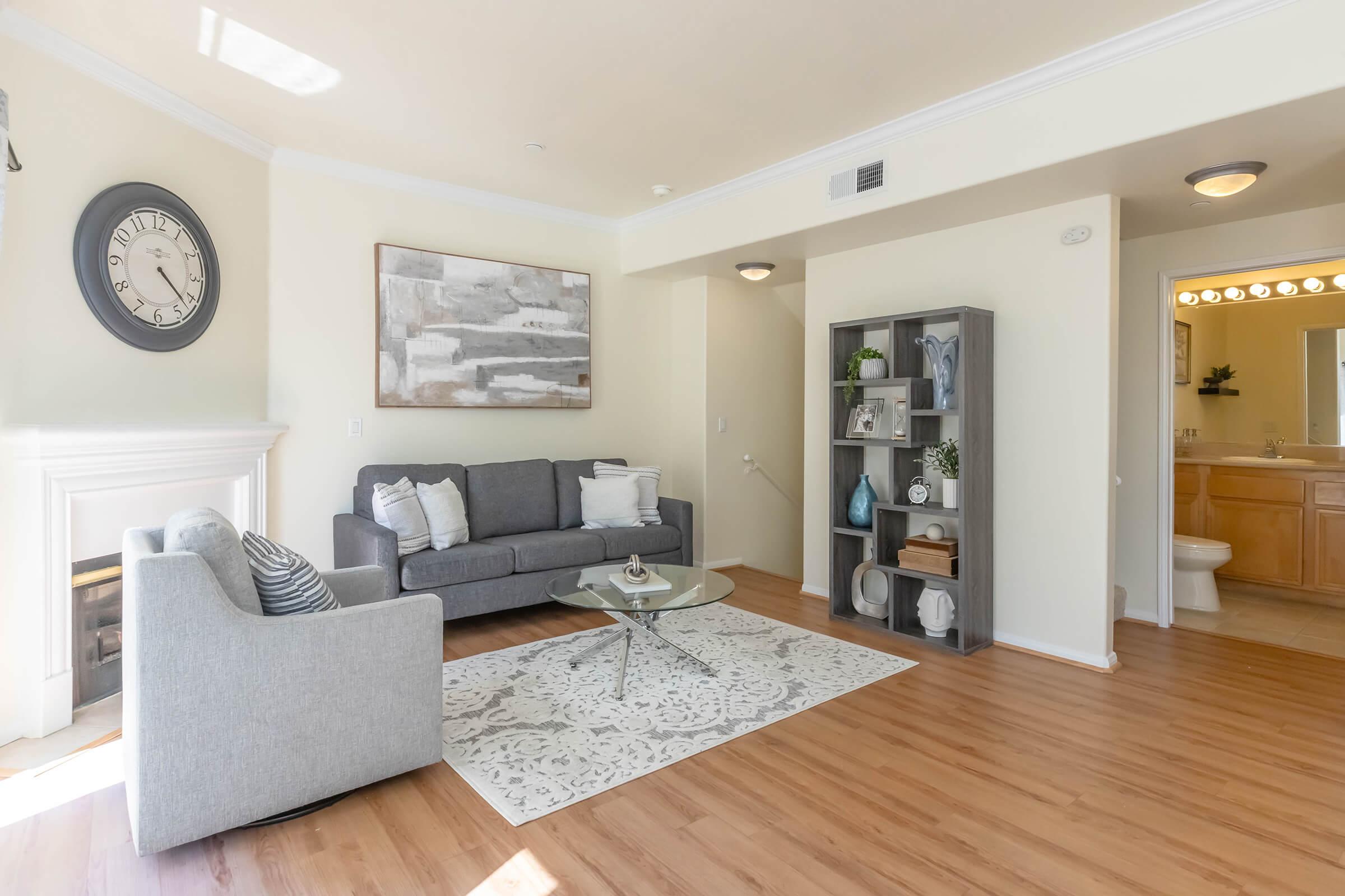 A bright living room featuring a gray sofa with white and gray cushions, a glass coffee table, and a decorative area rug. A framed painting hangs on the wall above the sofa, and a bookshelf with various decor items stands nearby. A doorway leading to a bathroom is visible in the background.