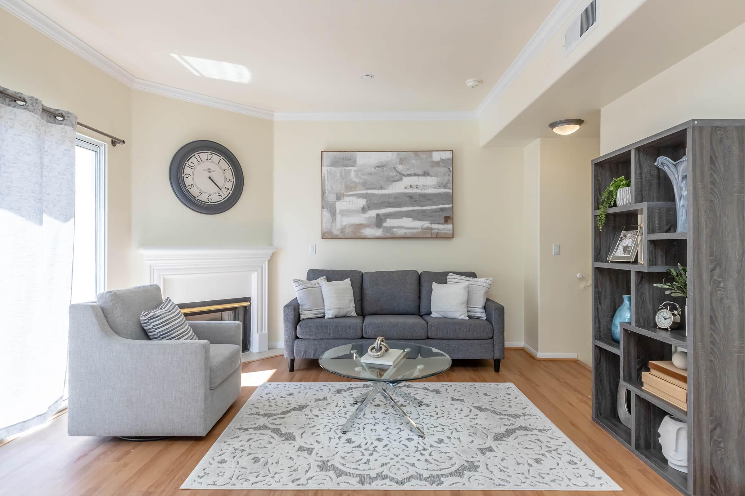 A modern living room featuring a gray sofa and an armchair, a circular glass coffee table on a patterned rug, and a wall clock. There’s a large window with sheer curtains allowing natural light, and a fireplace with decorative art above it. A bookshelf on the side holds various decorative items.