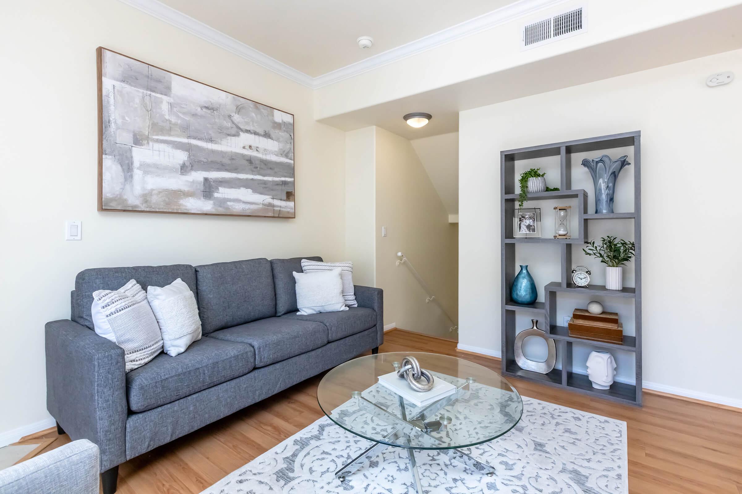 A modern living room featuring a gray couch with decorative pillows, a round glass coffee table on a light-colored rug, and a bookshelf filled with decorative items and plants. The walls are light-colored, and there's a staircase in the background. A large abstract painting hangs on one wall.