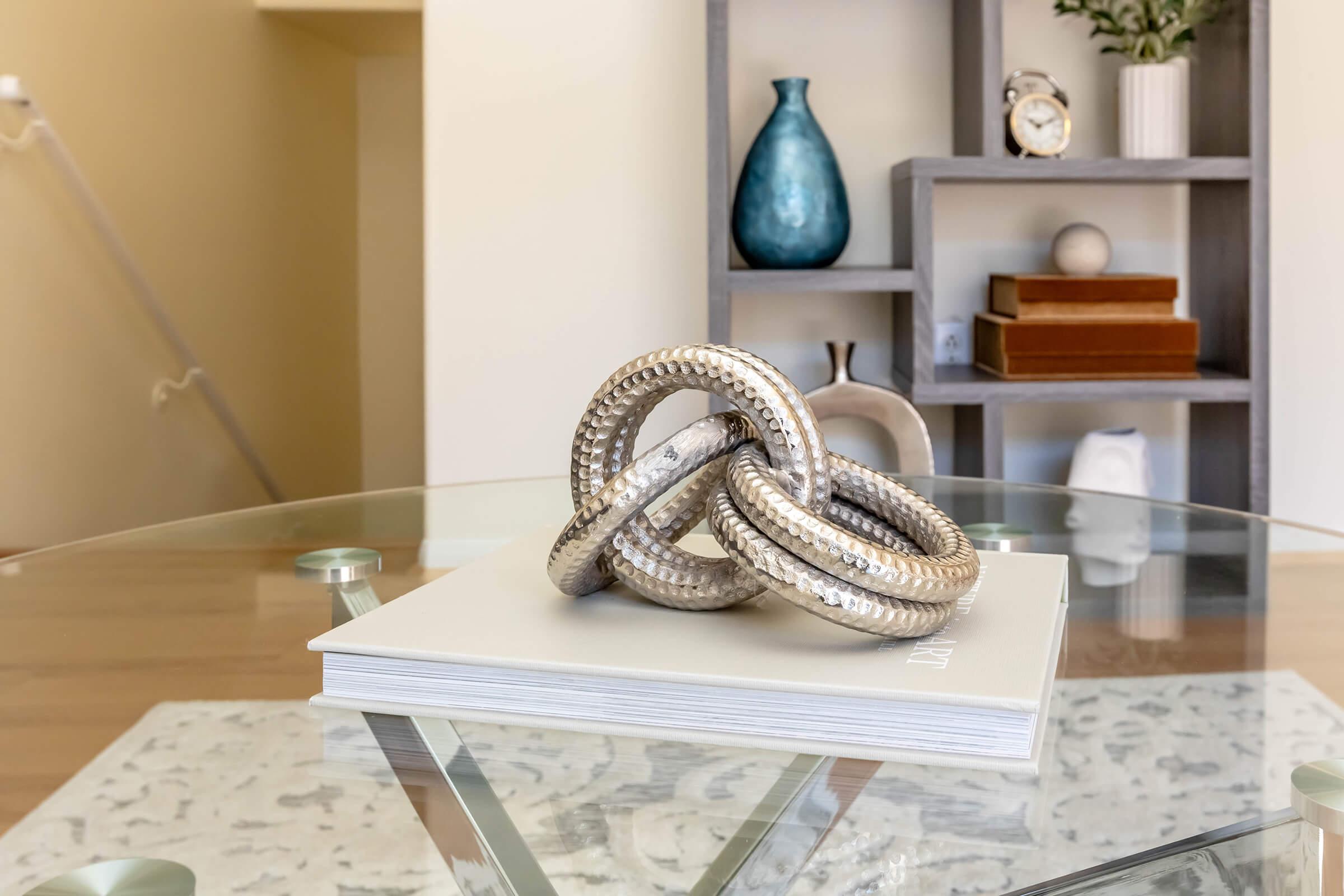A decorative arrangement of three intertwined metallic rings sits atop a coffee table book. In the background, there is a minimalist shelf displaying a vase, a clock, and some stacked boxes. The space features a warm and modern aesthetic with neutral colors.