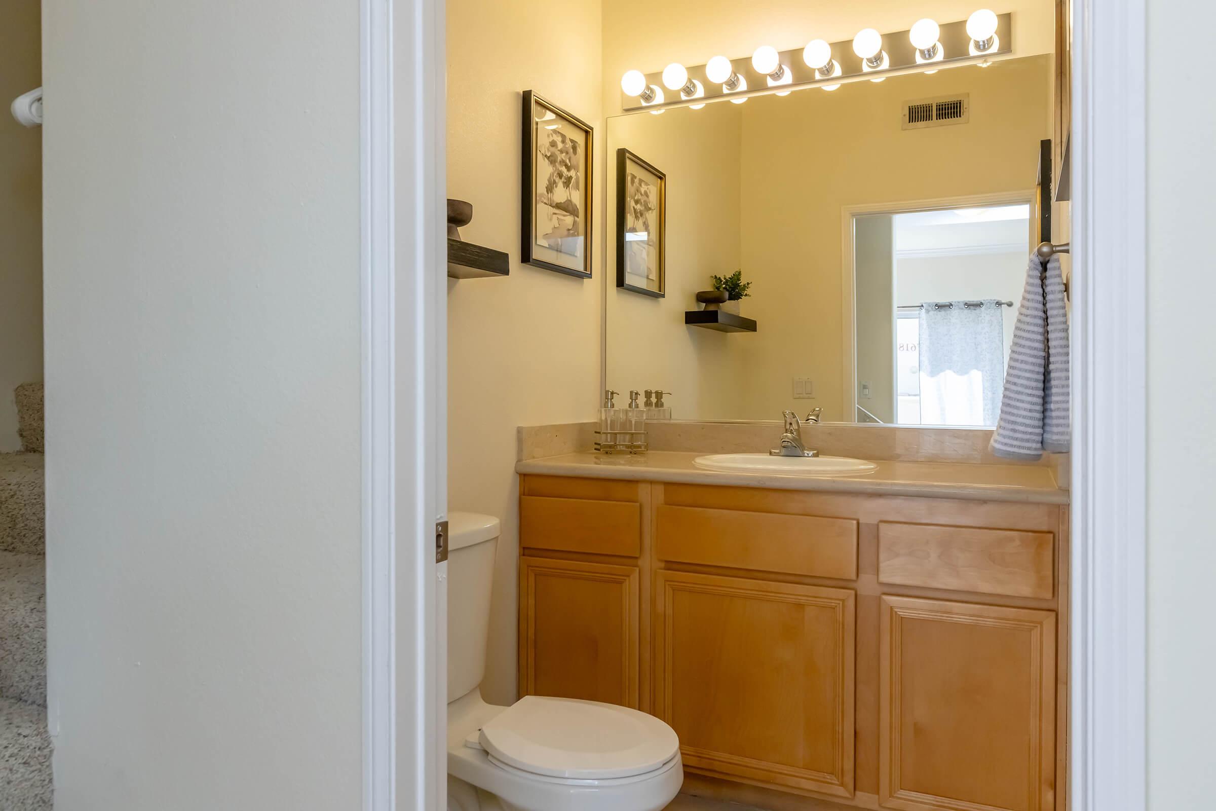 A well-lit bathroom featuring wooden cabinets, a marble countertop with a sink, and a large mirror. There are two framed pictures on the wall, decorative shelves, and a towel hanging. A toilet is visible, and stairs lead up to another area. The overall color scheme is light and inviting.