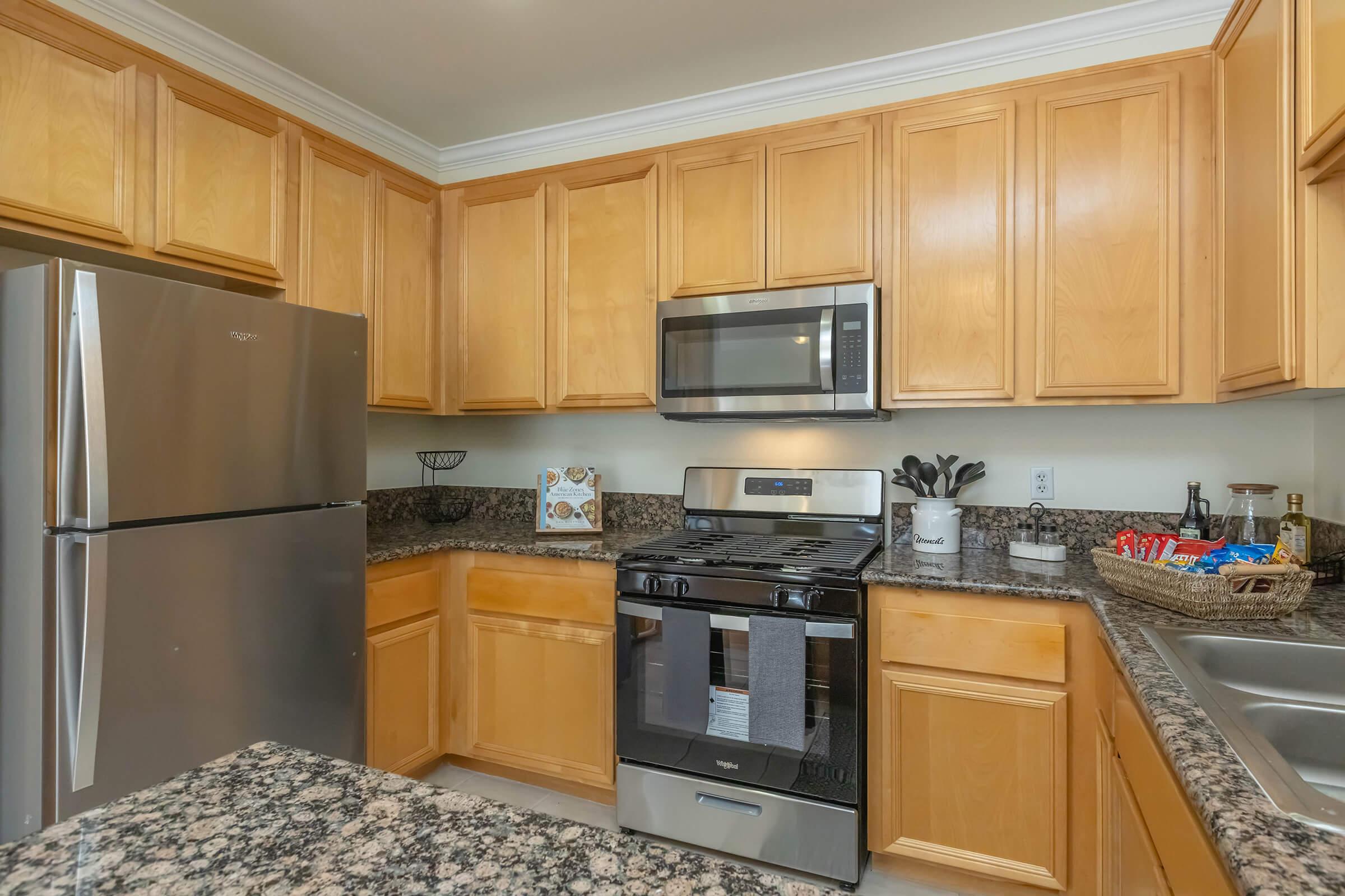 Modern kitchen featuring wooden cabinets, a stainless steel refrigerator, and a black stove with a microwave above. The countertops are made of granite, and there is a sink with utensils and a decorative basket on the side. The space is bright and well-organized.