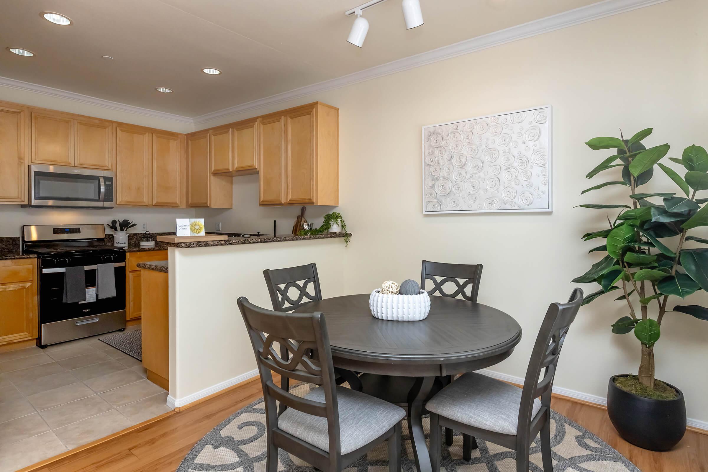 A cozy dining area featuring a round table with four chairs, a decorative centerpiece, and a rug. The kitchen, visible in the background, has wooden cabinets, stainless steel appliances, and a small decorative plant. A framed wall art piece adds a touch of decor, and a potted plant is positioned in the corner.