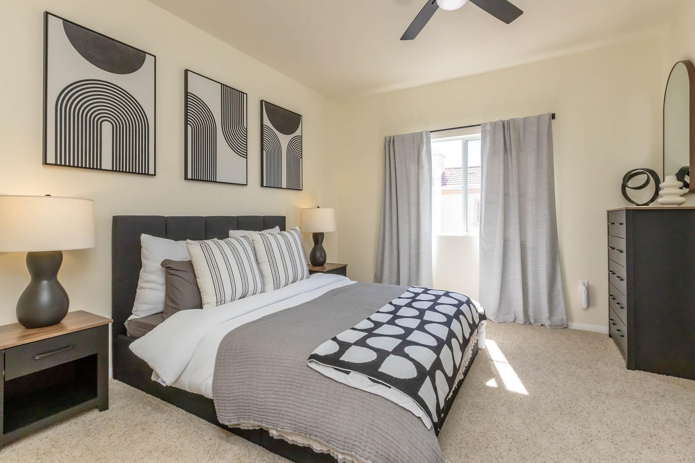 A modern bedroom featuring a large bed with gray and white bedding, two bedside tables with lamps, and framed abstract artwork on the walls. Light curtains cover a window, allowing natural light to brighten the space, which includes a dresser and plush carpet flooring.