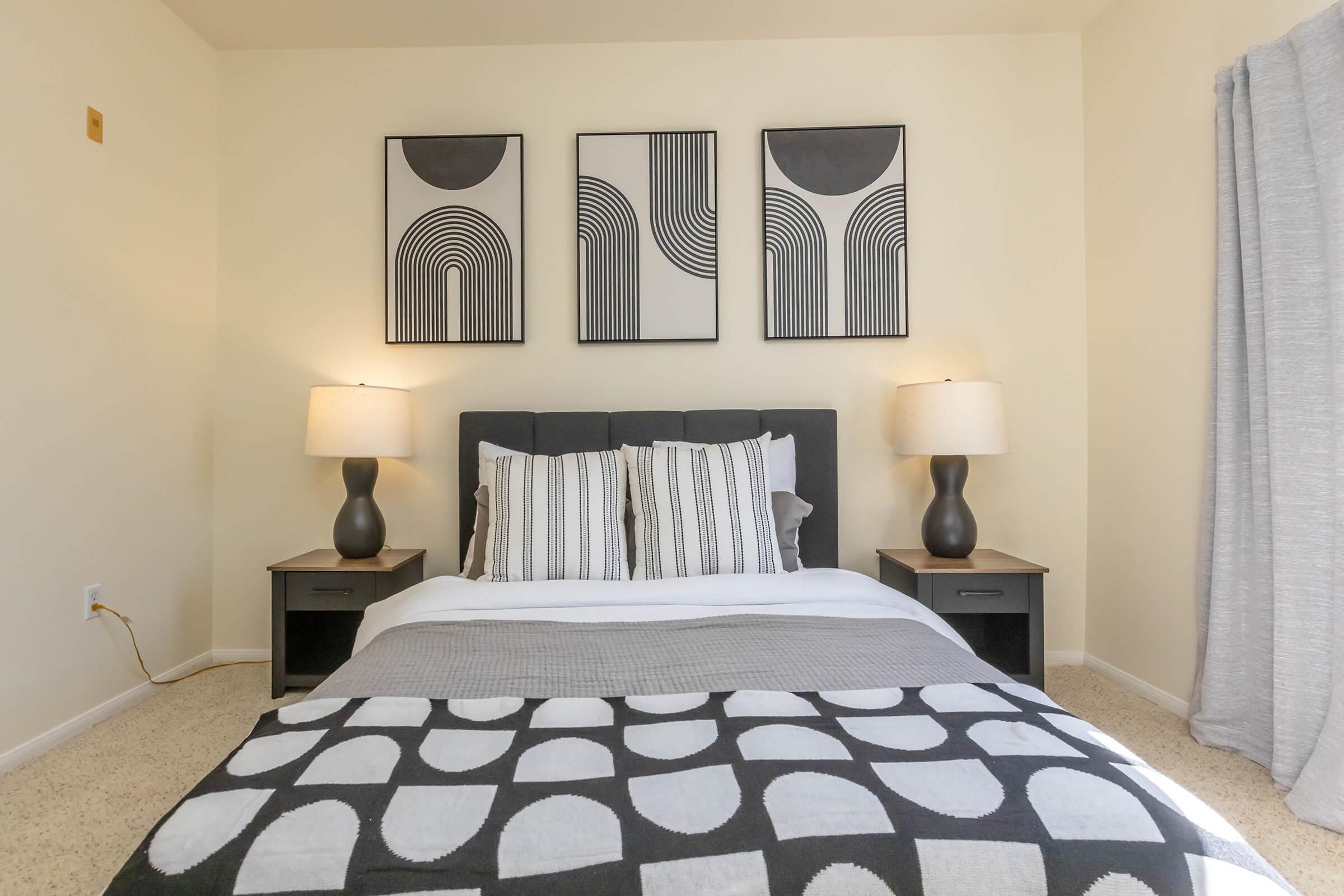 A modern bedroom featuring a queen-sized bed with gray and white bedding. There are two bedside tables with black lamps on each side. Above the bed are three geometric wall art pieces in black and white. The room has light beige walls and a soft-textured gray curtain covering a window.